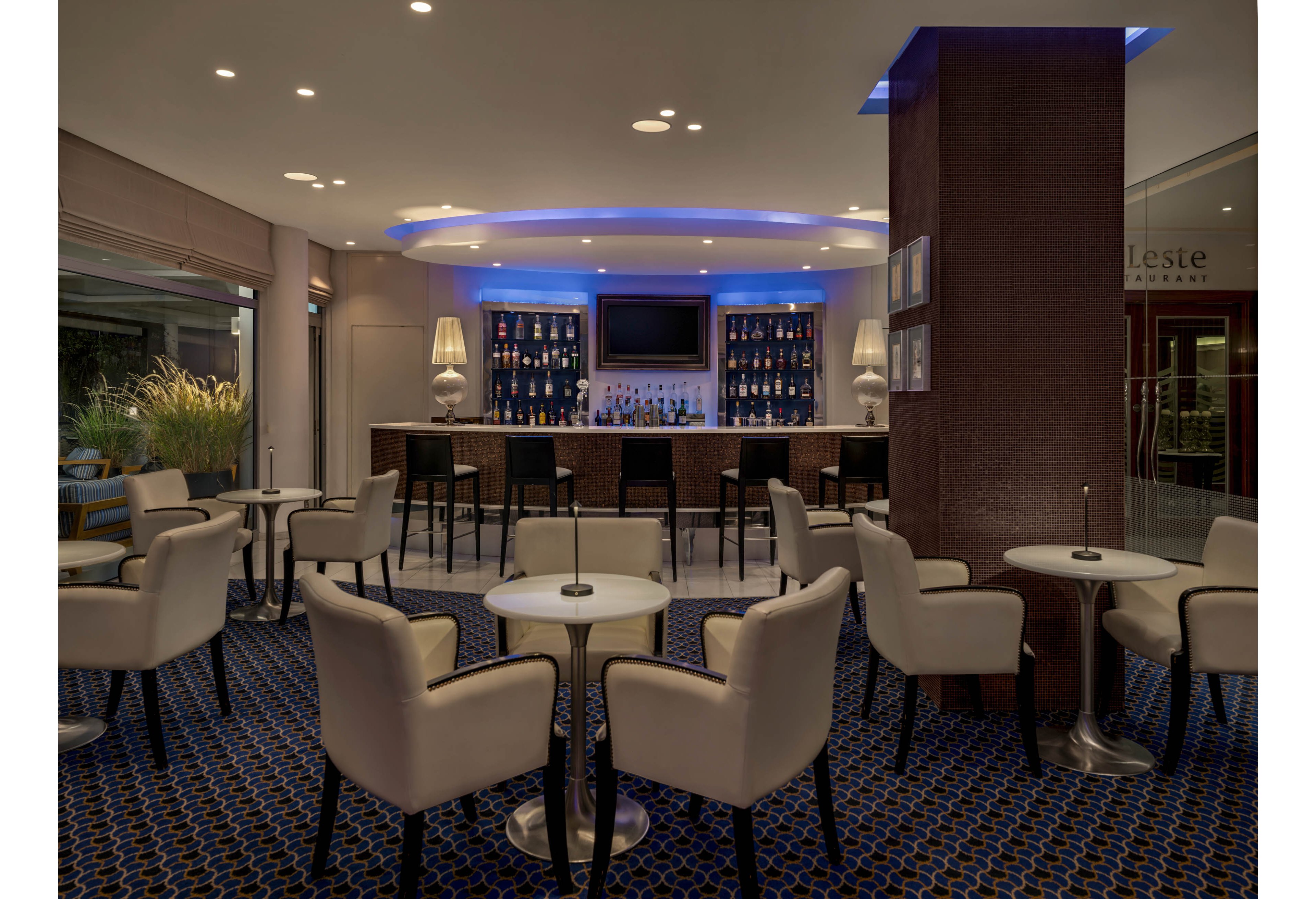 A contemporary hotel lounge bar features a sleek counter lined with barstools and a backlit display of bottles. Soft blue accent lighting highlights the ceiling and bar shelves, creating a sophisticated ambiance. White upholstered armchairs and small round tables are arranged neatly across a patterned carpet. Large windows reveal an outdoor area with plants, adding a touch of greenery to the interior.