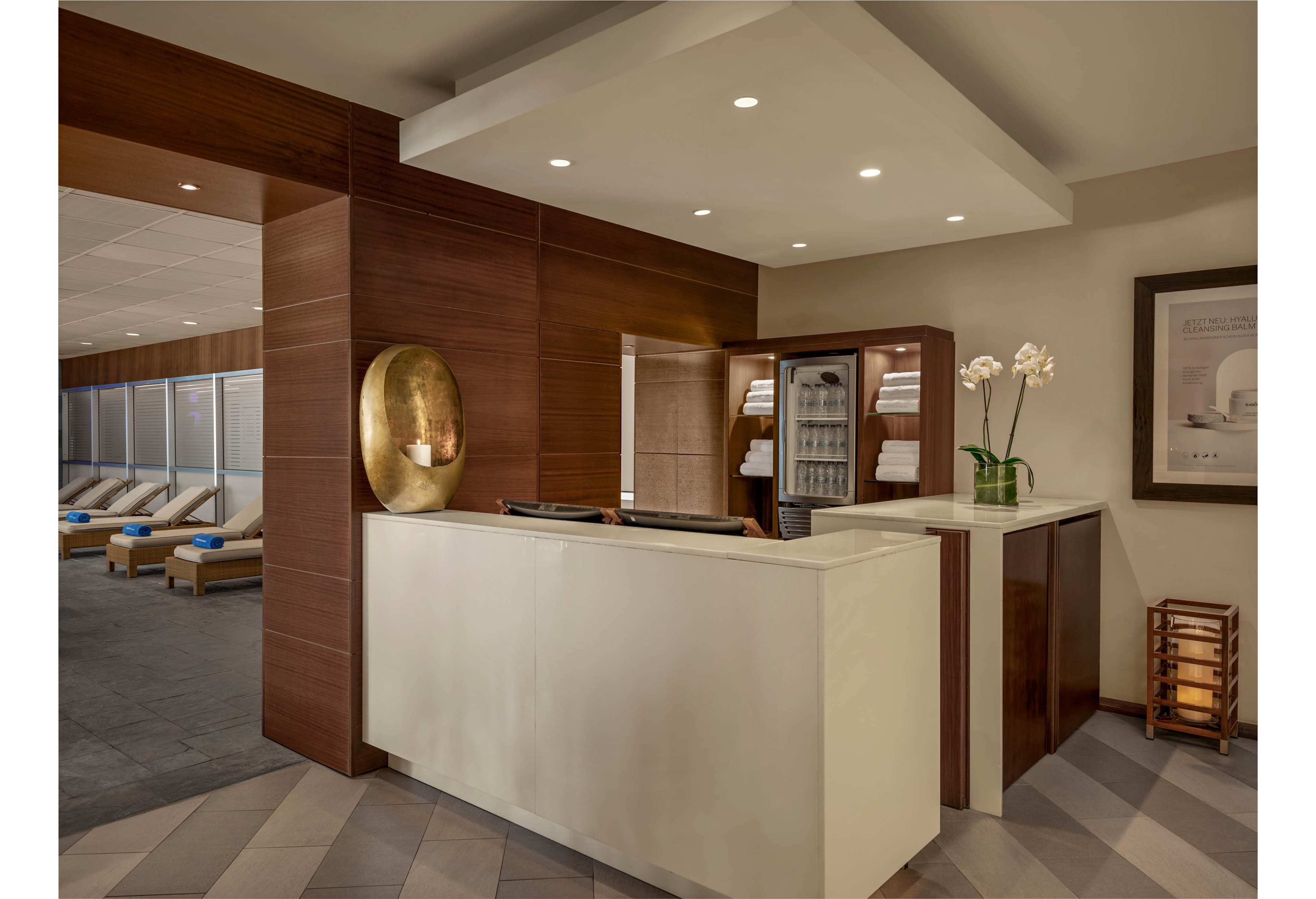 A contemporary spa reception area features a sleek white front desk framed by rich wooden paneling. Behind the counter, neatly folded towels are stacked in open shelving, suggesting a wellness or relaxation setting. Soft recessed lighting and neutral tones create a calm, inviting atmosphere. In the background, lounge chairs with blue cushions line the wall, while decorative elements like orchids and wall art add a refined touch.