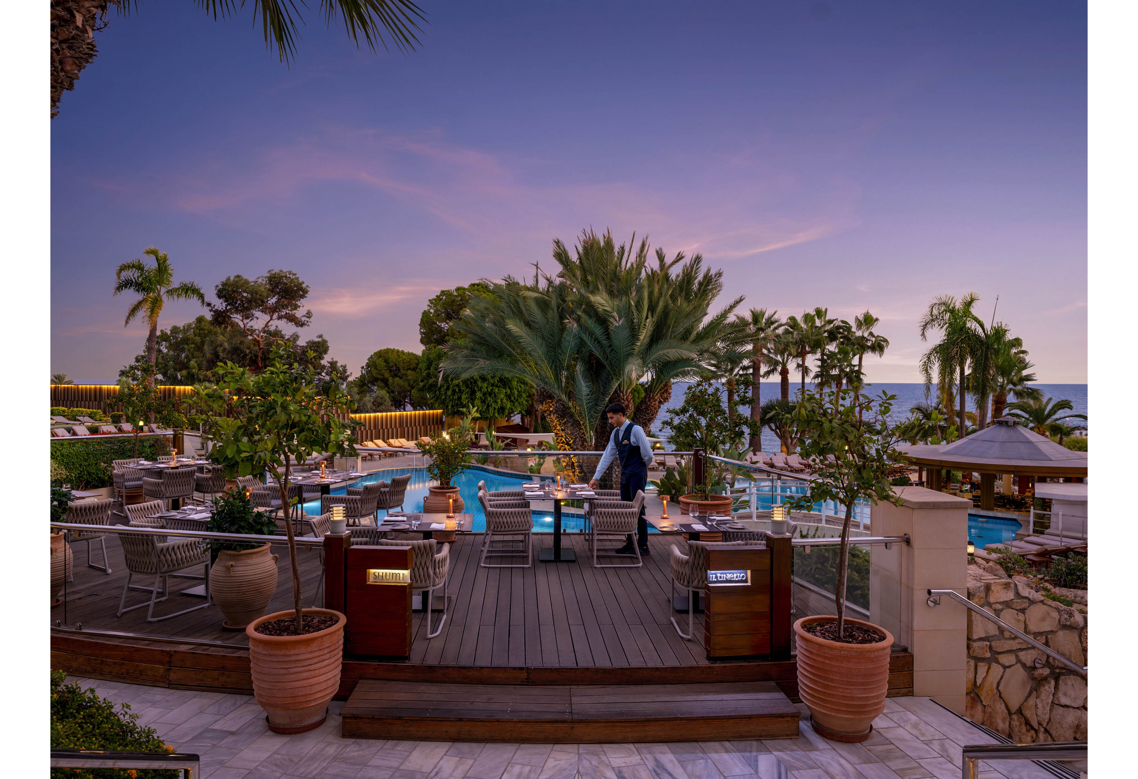 An upscale outdoor restaurant overlooks a resort pool at sunset. A server stands on the wooden deck among neatly arranged tables and chairs, preparing the space for guests. Lush palm trees, potted plants, and soft lighting create a tropical, coastal ambiance. The calm sea and pastel evening sky enhance the relaxed, luxurious atmosphere.