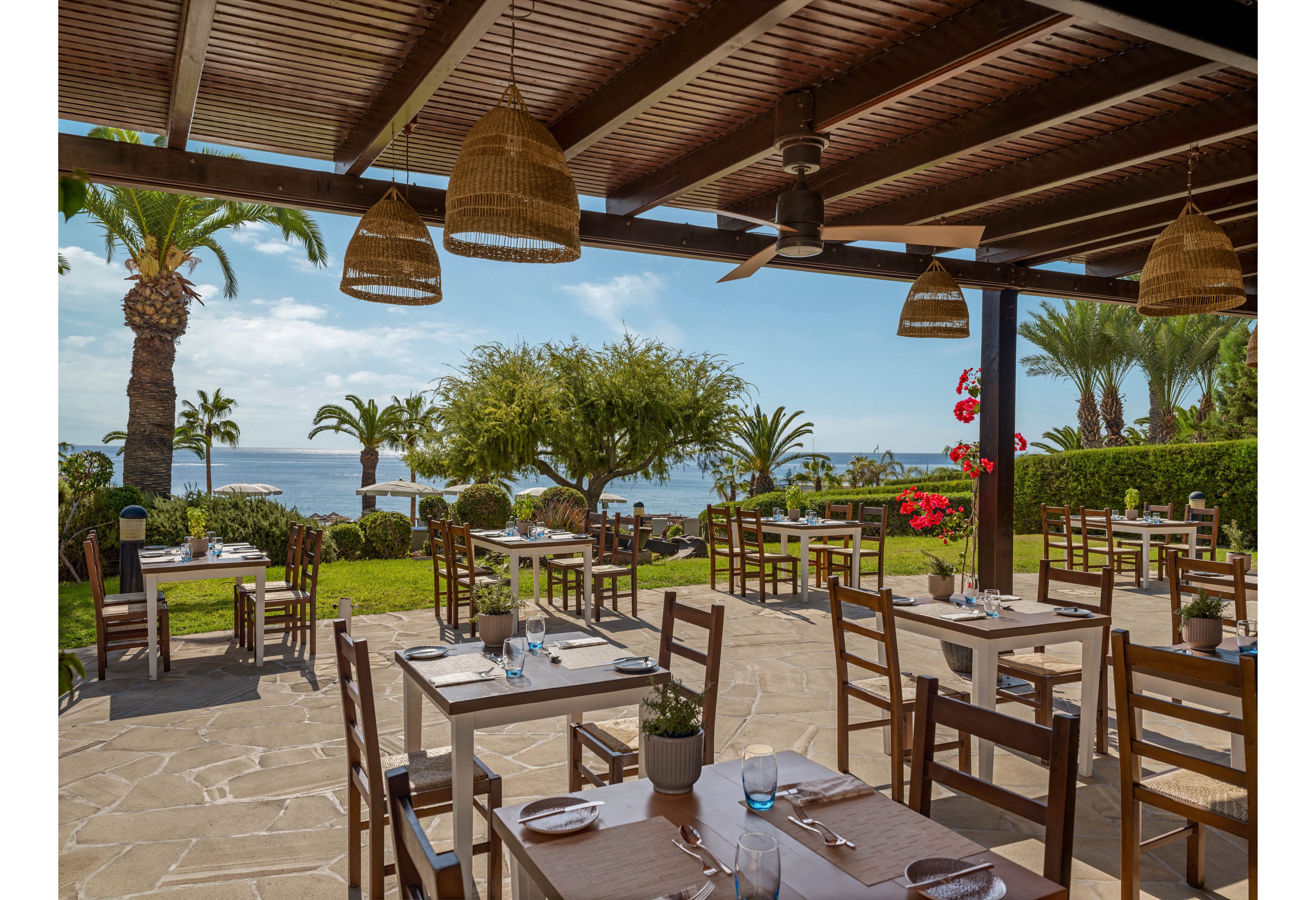 An open-air restaurant terrace overlooks a calm seaside landscape with palm trees and lush greenery. Wooden tables and chairs are neatly arranged under a shaded pergola, each set with plates, cutlery, and glasses. The bright daylight highlights the blue sea in the background and colorful flowers along the garden edge. The scene feels relaxed and inviting, ideal for outdoor dining by the water.