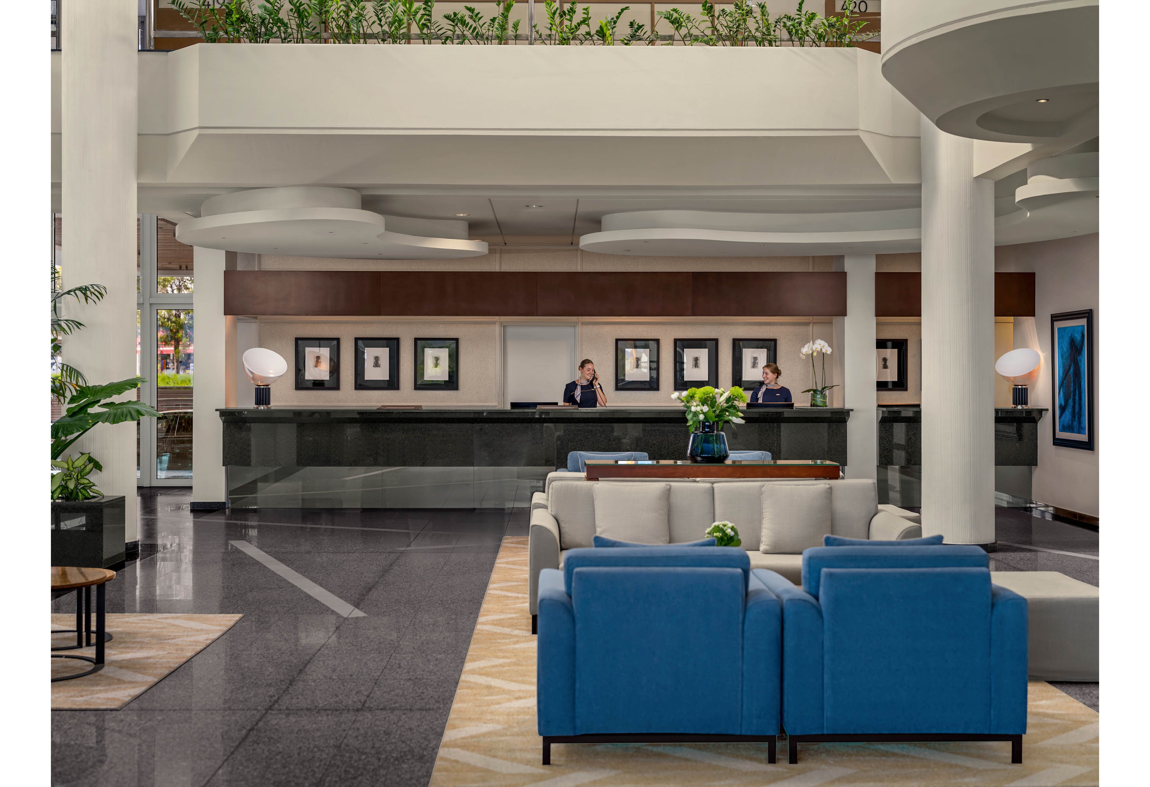 A contemporary hotel lobby features a sleek reception desk staffed by two people. In the foreground, blue and beige sofas create a comfortable seating area with a central table and floral arrangement. The space has polished dark flooring, white columns, and layered ceiling details that add architectural interest. Framed artwork and soft lighting contribute to a welcoming, upscale atmosphere.