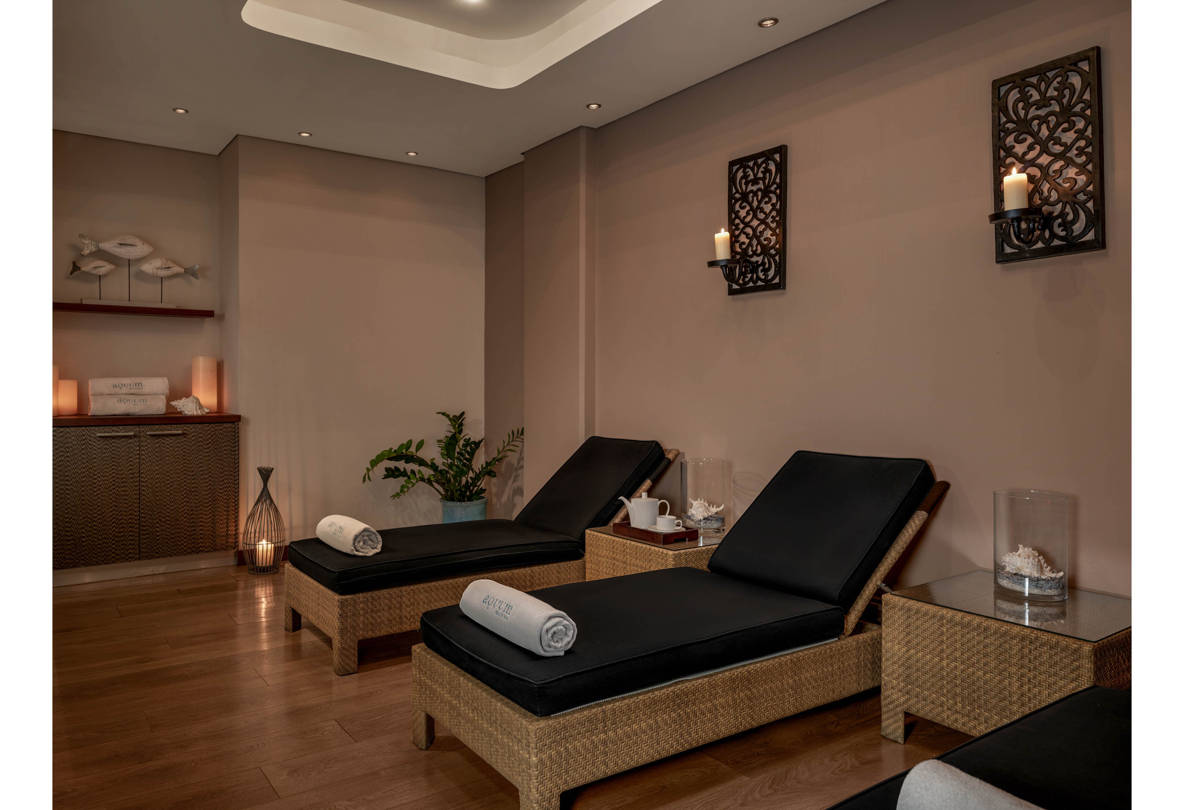 A tranquil spa lounge features wicker reclining chairs with black cushions and neatly rolled white towels. Soft candlelight glows from wall sconces and decorative lanterns, creating a warm and relaxing atmosphere. A potted plant and minimalist wall art add natural and artistic touches to the neutral-toned room. The wooden floor and subtle lighting emphasize comfort and calm, suggesting a space for rest and wellness treatments.