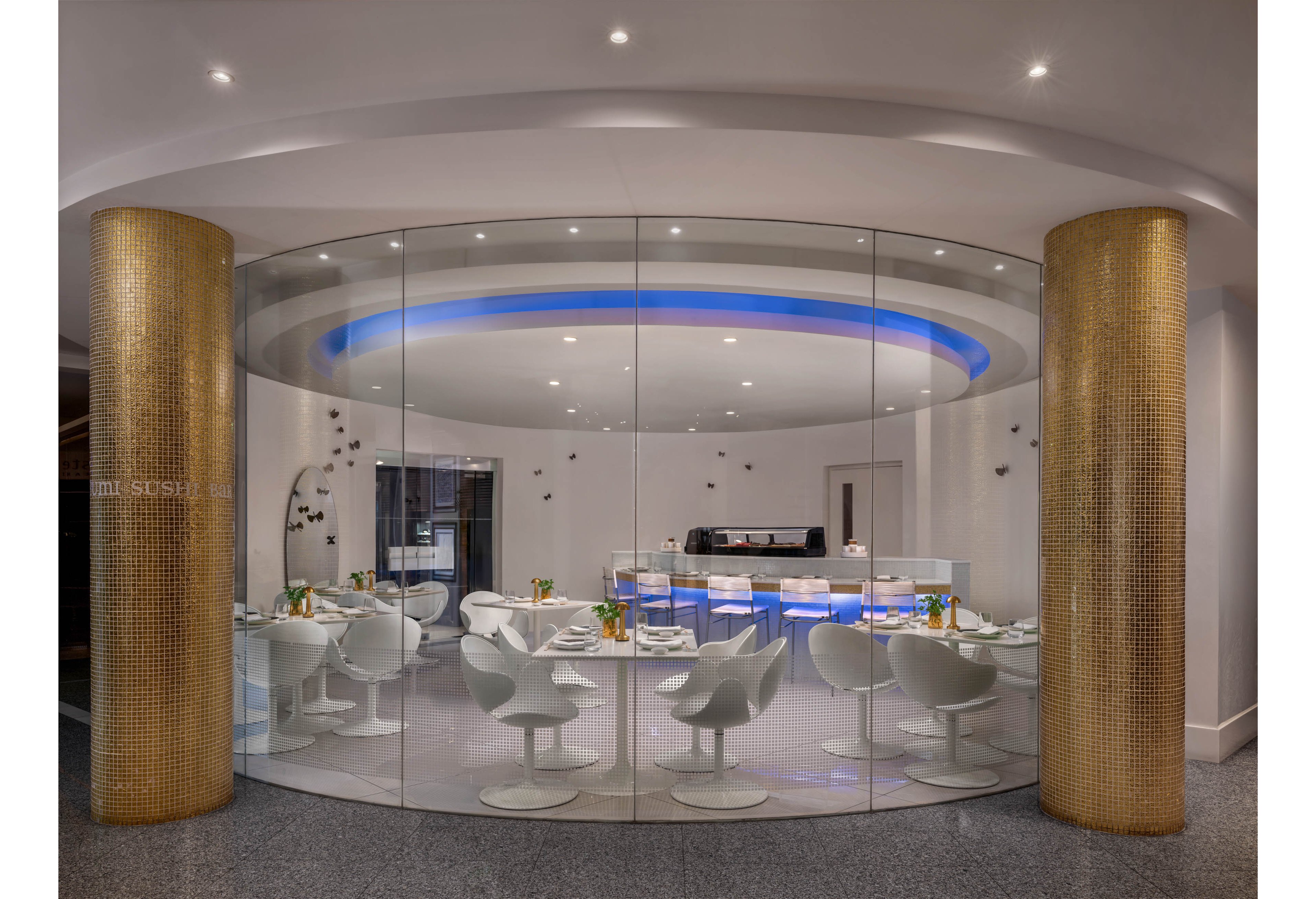 A contemporary circular dining room is enclosed by floor-to-ceiling glass walls and framed by two golden cylindrical columns. Inside, white modern chairs and round tables are neatly arranged, set with plates, glasses, and small floral accents. A softly glowing blue light ring highlights the ceiling above the central bar or counter area. The overall design feels sleek and futuristic, with neutral tones accented by metallic and blue lighting details.