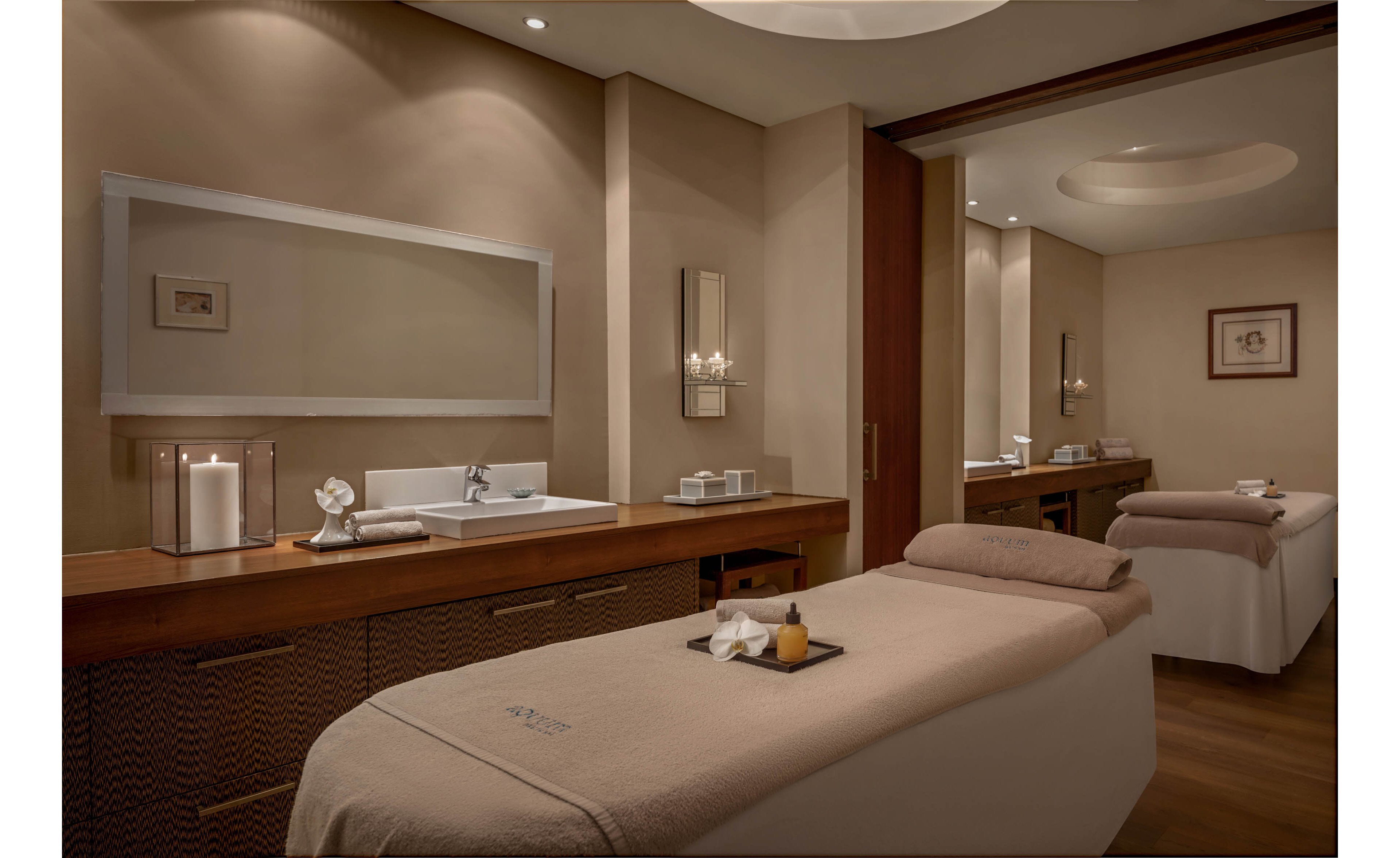 A serene spa treatment room features two neatly prepared massage tables with folded towels and small trays. Soft, warm lighting and neutral beige tones create a calming atmosphere. A long wooden counter with a sink, candles, and decorative items lines one wall, while framed artwork adds subtle detail. The space appears quiet and inviting, ideal for relaxation and wellness treatments.