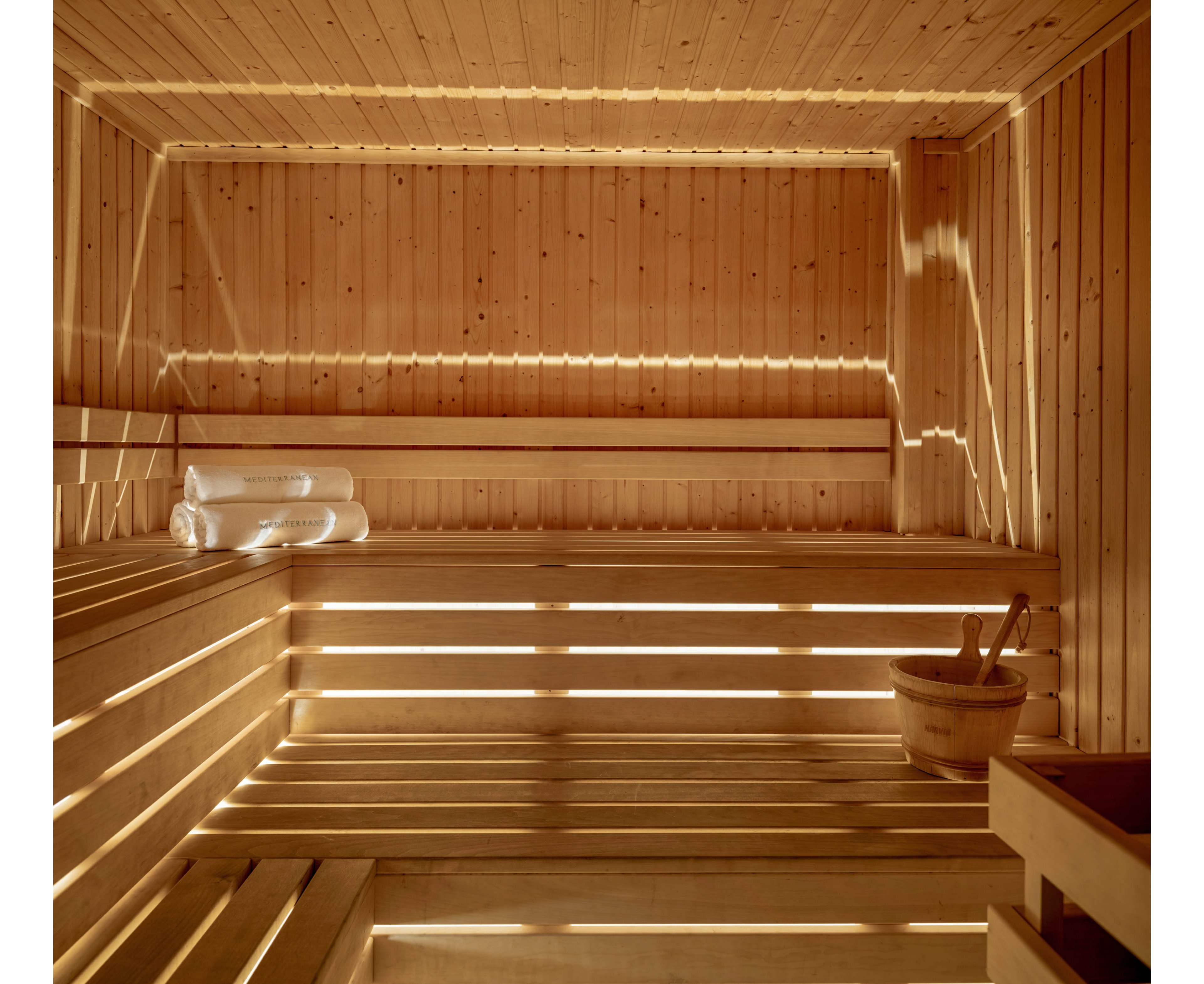 A serene wooden sauna interior features tiered benches and soft, warm lighting. Sunlight filters through narrow slats, creating linear patterns across the walls and seating. A wooden bucket with ladle sits on a bench, suggesting a traditional steam sauna experience. Rolled white towels are neatly stacked, enhancing the spa-like, relaxing atmosphere.
