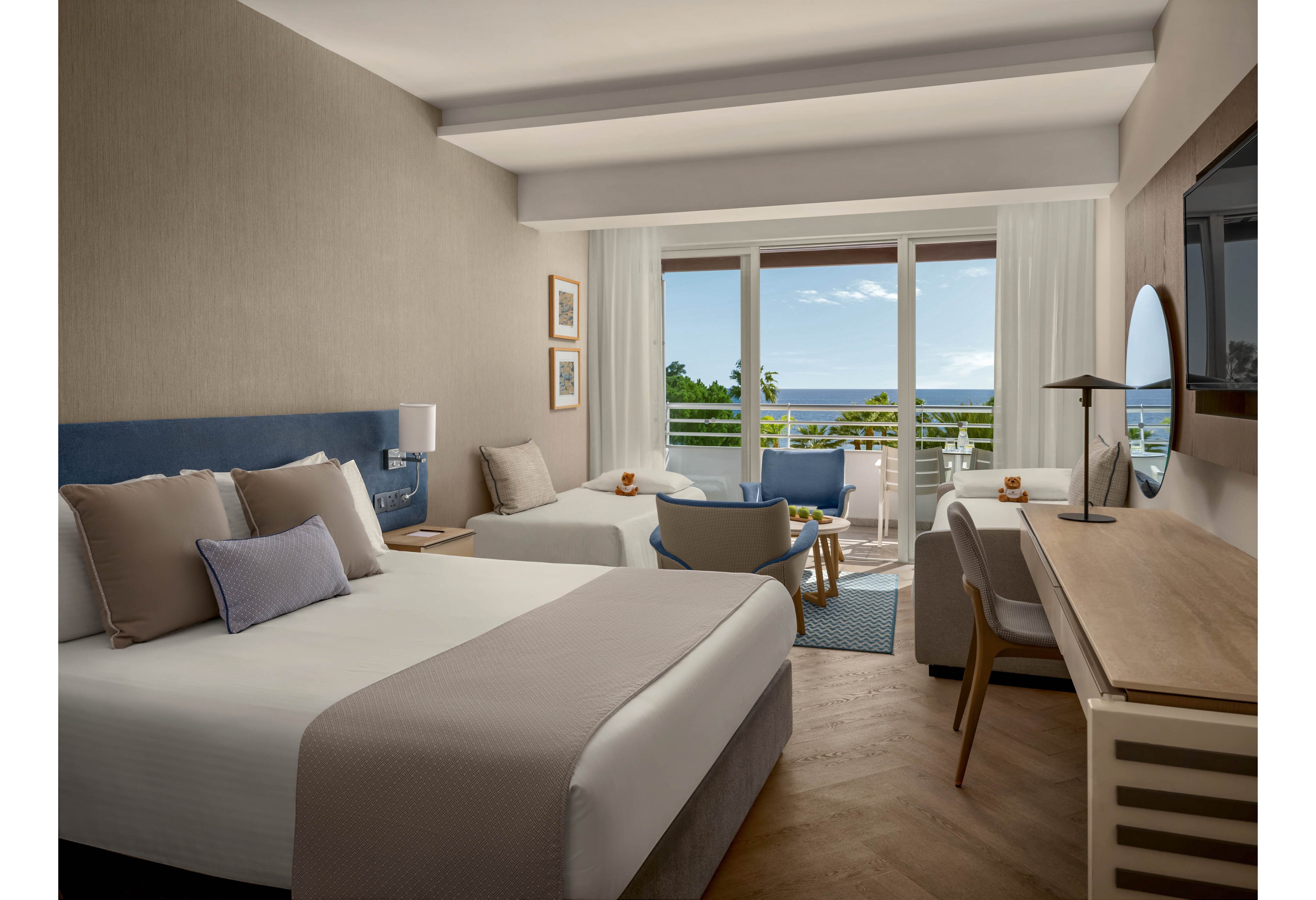A contemporary hotel room features a large bed and a smaller bed arranged toward a balcony. Neutral tones of beige, white, and soft blue create a calm and inviting atmosphere. A desk with a chair, table lamp, and wall-mounted television line one side of the room, while a seating area with armchairs sits near the window. Floor-to-ceiling curtains frame the balcony doors, revealing a view of trees and the distant ocean under a clear sky.