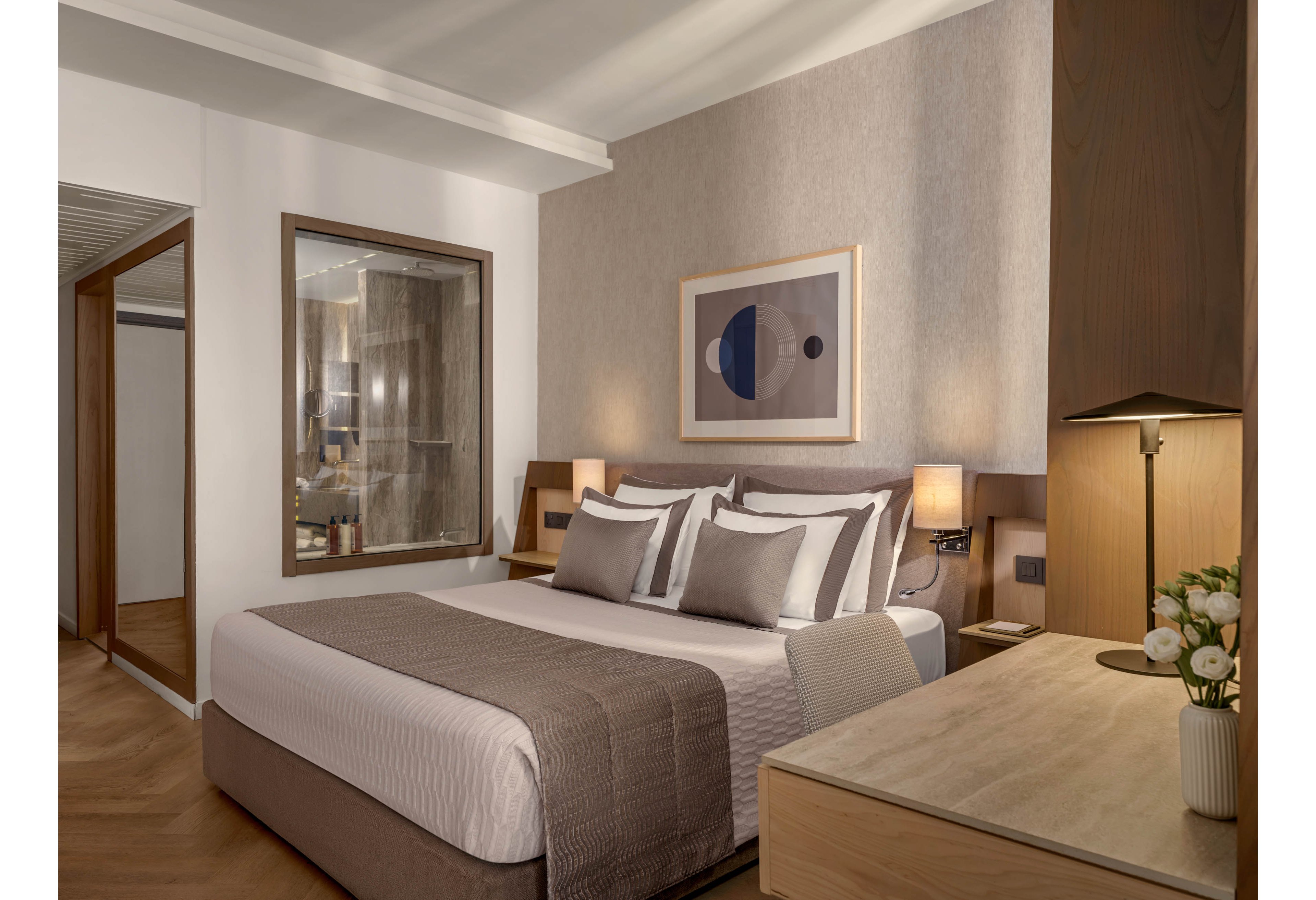 A contemporary hotel bedroom features a neatly made double bed with layered pillows and neutral-toned bedding. The room includes a wooden desk with a small vase of flowers and warm bedside lighting. A large glass window reveals the adjoining bathroom, adding a sense of openness to the space. Minimalist artwork and soft beige and brown tones create a calm, sophisticated atmosphere.