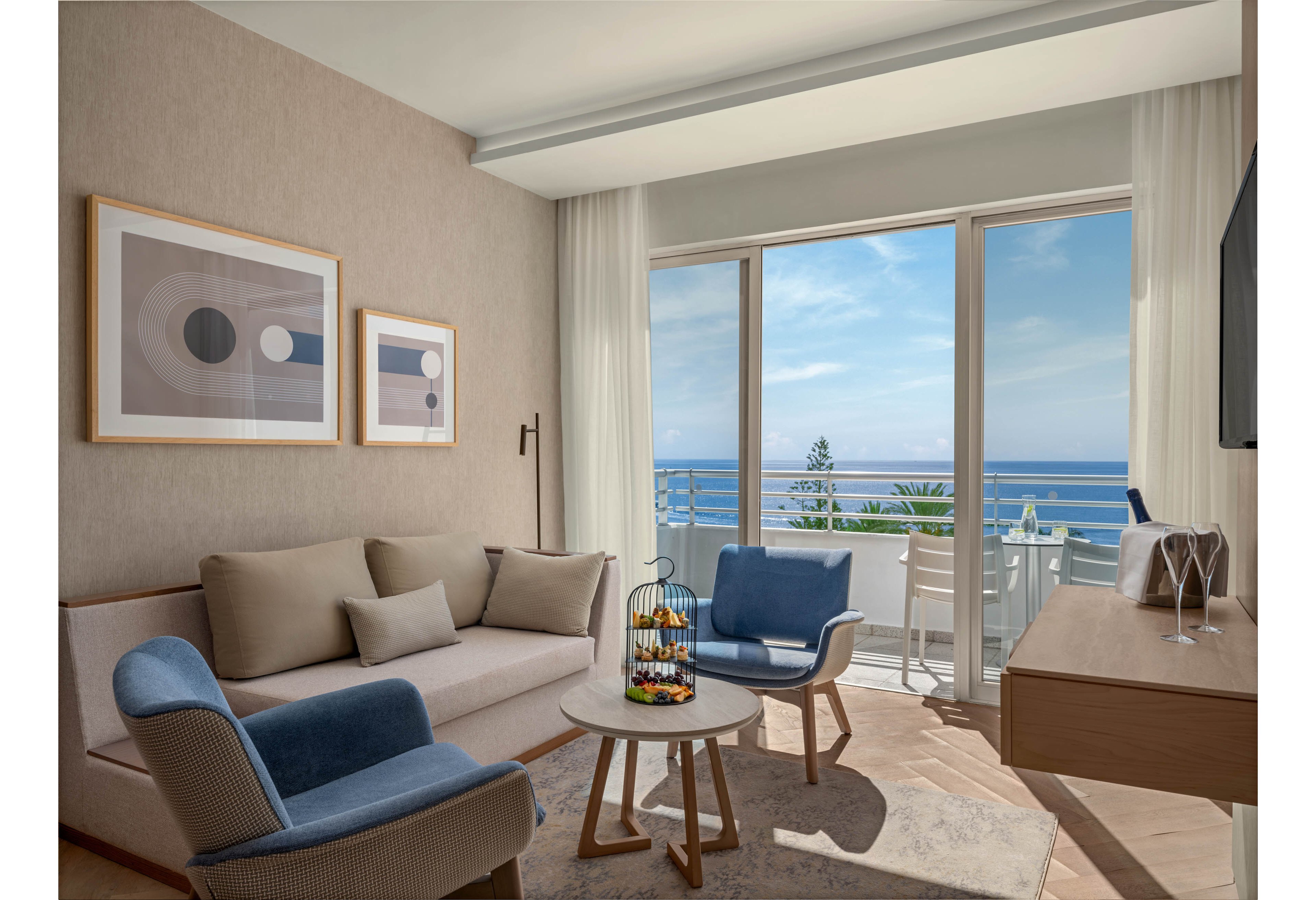 A contemporary hotel living room features a neutral-toned sofa and armchairs arranged around a small round coffee table. Large sliding glass doors open to a balcony with white outdoor furniture and a clear view of the ocean and blue sky. Minimalist abstract artwork decorates the wall, and sheer curtains frame the bright natural light. The space feels calm and airy, combining soft beige and blue accents with clean modern lines.