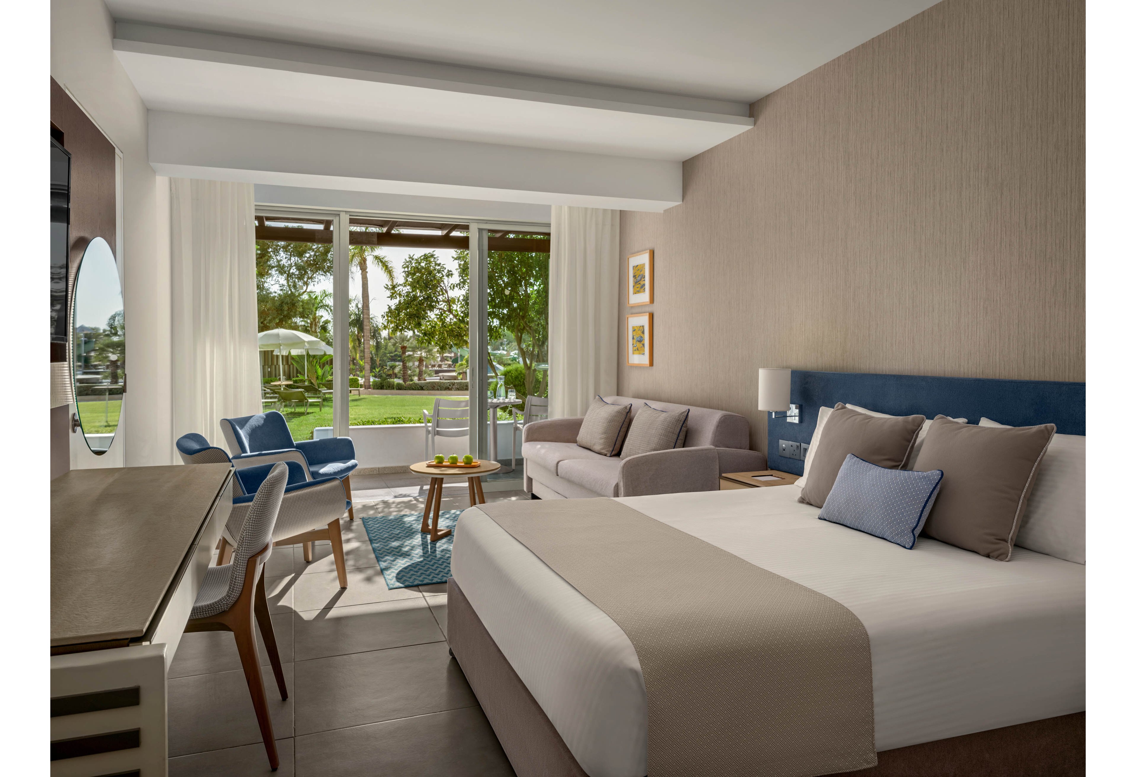 A contemporary hotel room features a neatly made double bed with neutral bedding and accent pillows. A sofa, armchair, and small coffee table create a cozy seating area near the window. Large glass doors open to a terrace with outdoor chairs and a view of a green lawn and trees. Soft natural light, minimalist furniture, and muted colors give the space a calm, relaxing atmosphere.