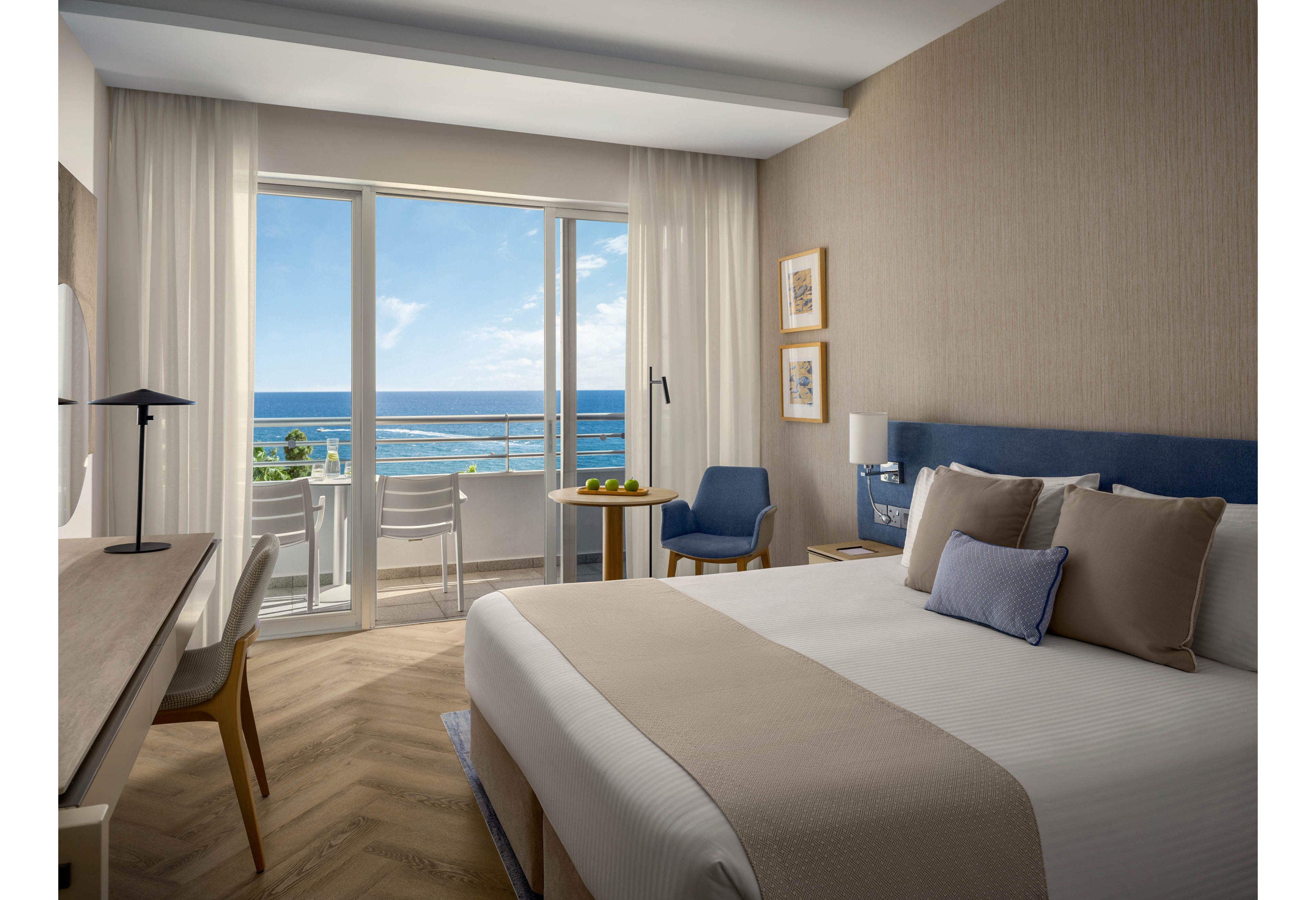 A contemporary hotel bedroom features a neatly made double bed with neutral-toned linens and a padded headboard. Large glass doors open onto a balcony with white chairs and a table, overlooking a bright blue ocean and clear sky. The room includes a desk with a lamp, a small round table with fruit, and a blue accent chair. Soft natural light and beige walls create a calm, inviting atmosphere ideal for relaxation.