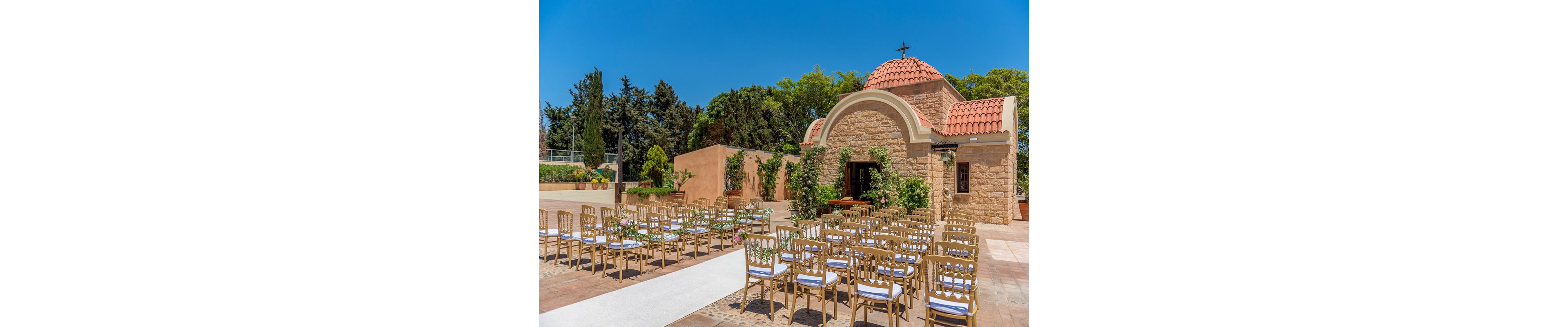 Rows of elegant gold chairs are arranged for an outdoor wedding ceremony in front of a small stone chapel. The chapel features a terracotta-tiled dome, arched doorway, and a cross on top, surrounded by lush greenery. A white aisle runner leads toward the entrance, decorated with floral arrangements on the chairs. The scene is brightly lit under a clear blue sky, creating a festive and romantic atmosphere.
