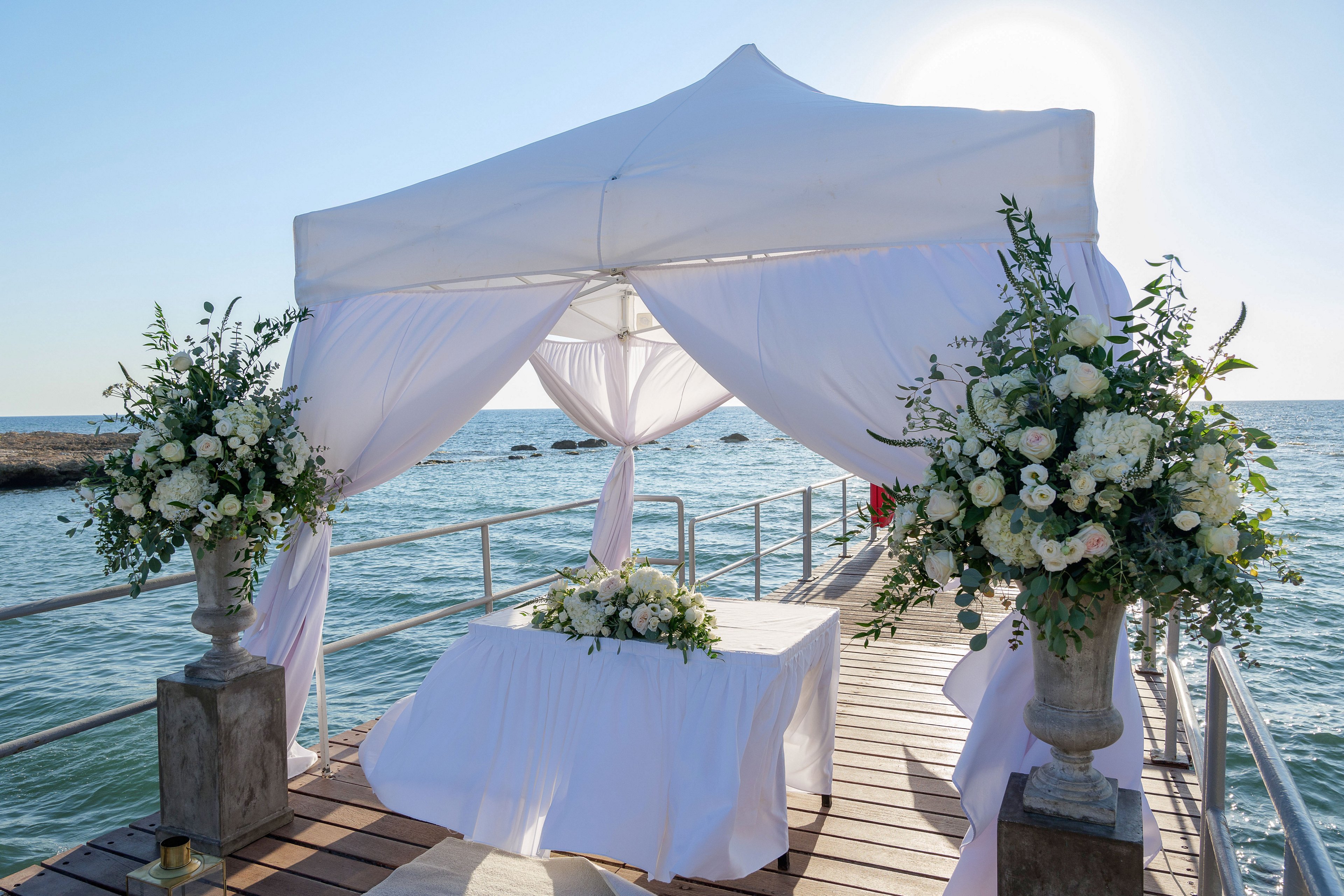 A white canopy wedding altar is set up on a wooden pier extending over the sea. Flowing white drapes frame the structure, creating a light and airy look against the blue water and sky. Large floral arrangements with white flowers and greenery stand on both sides of the entrance, with a matching arrangement on a central table. The scene is bright and serene, suggesting a romantic outdoor ceremony at the water’s edge.