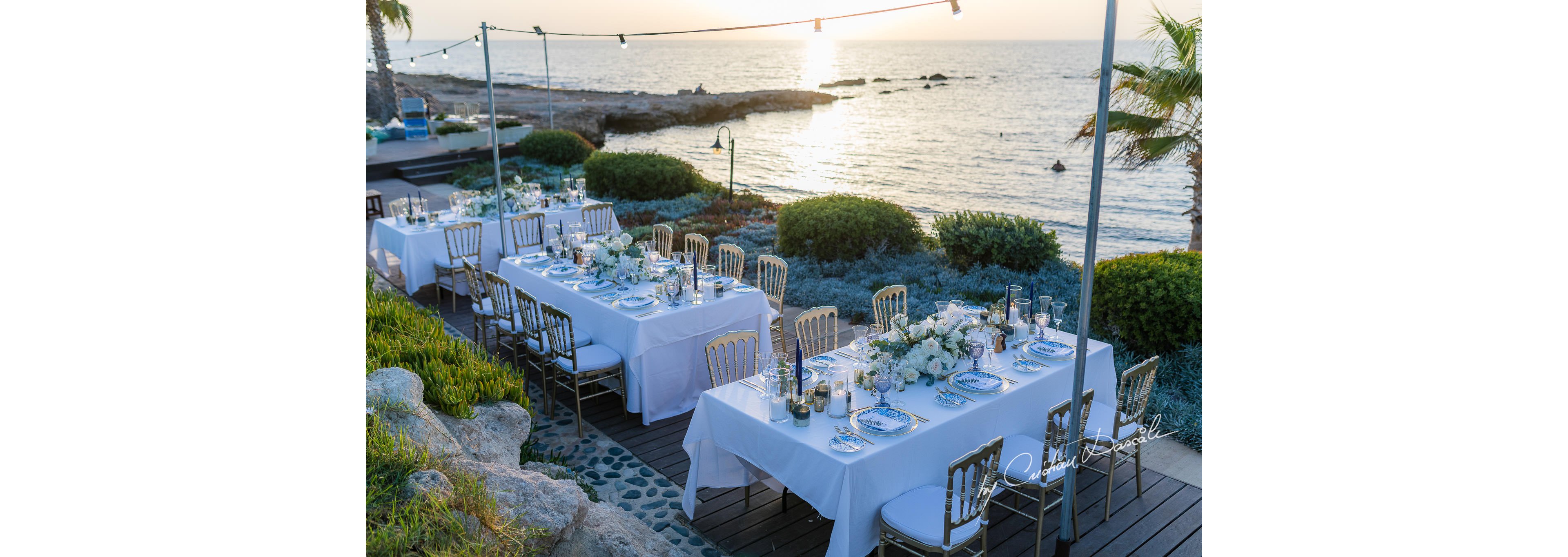An elegant outdoor reception is set up on a wooden deck overlooking the sea at sunset. Long white-clothed tables are arranged with floral centerpieces, candles, and neatly arranged place settings. Gold and white chairs line the tables, creating a refined and romantic atmosphere. String lights are suspended above the dining area, while the rocky shoreline and calm water provide a scenic coastal backdrop.