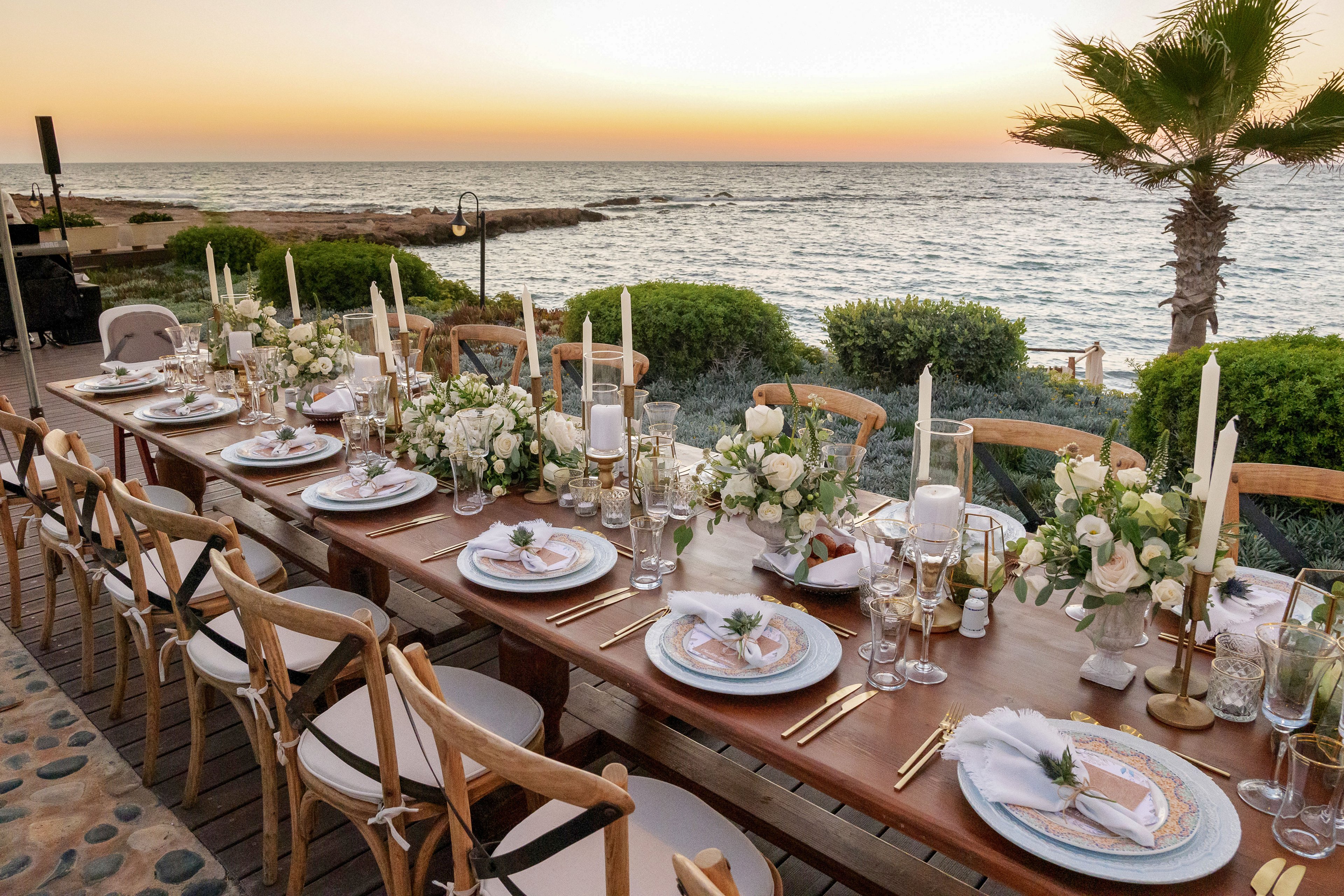 A long wooden dining table is beautifully set for an outdoor event beside the sea at sunset. White plates, linen napkins, and gold cutlery are arranged with tall candles and glassware. Lush white floral centerpieces and greenery run along the center of the table, complementing the coastal landscape. Wooden chairs line both sides of the table, creating an inviting atmosphere for a formal celebration by the water.