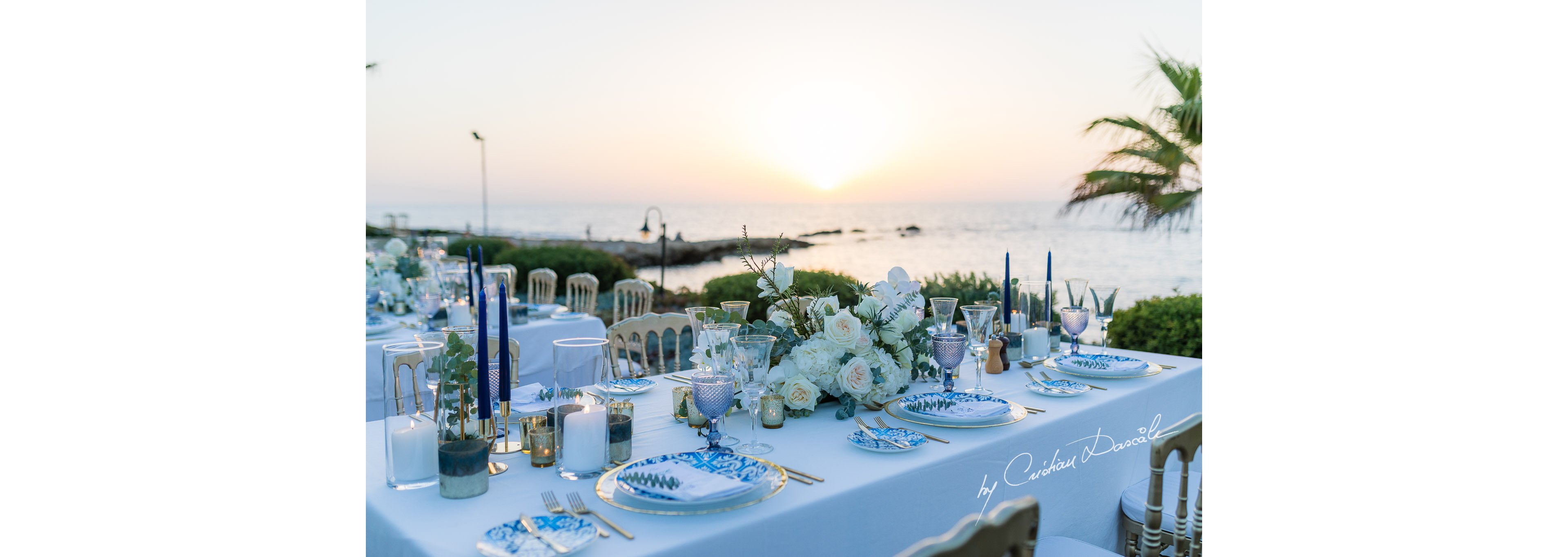 A long, elegantly set dining table is arranged outdoors beside the sea at sunset. White linens, blue-patterned plates, and tall blue candles create a refined coastal theme. A large white floral centerpiece with greenery decorates the center of the table, surrounded by glassware and small votive candles. The calm ocean, palm trees, and warm evening light provide a romantic backdrop for the celebration.