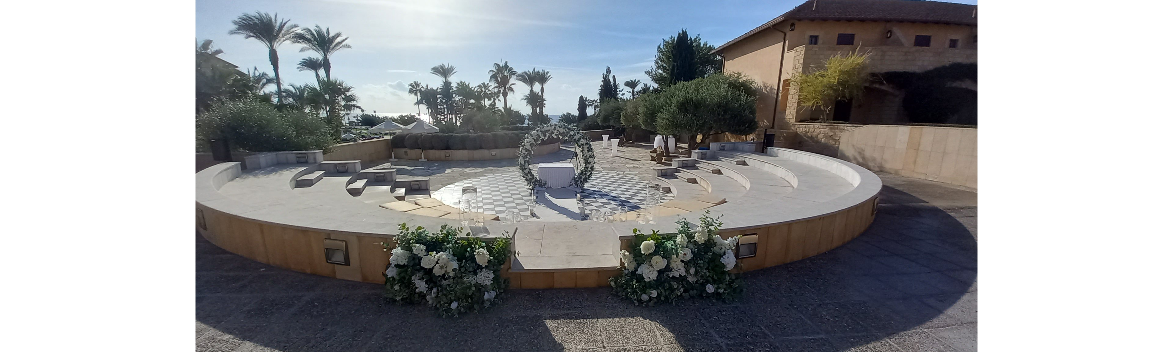 An elegant circular wedding ceremony stage is arranged in an outdoor courtyard. A white aisle leads to a central table framed by a floral arch and surrounded by curved seating steps. Lush white flower arrangements decorate the front of the platform, while palm trees and greenery create a scenic backdrop. Warm sunlight and clear skies enhance the romantic, Mediterranean-style atmosphere.