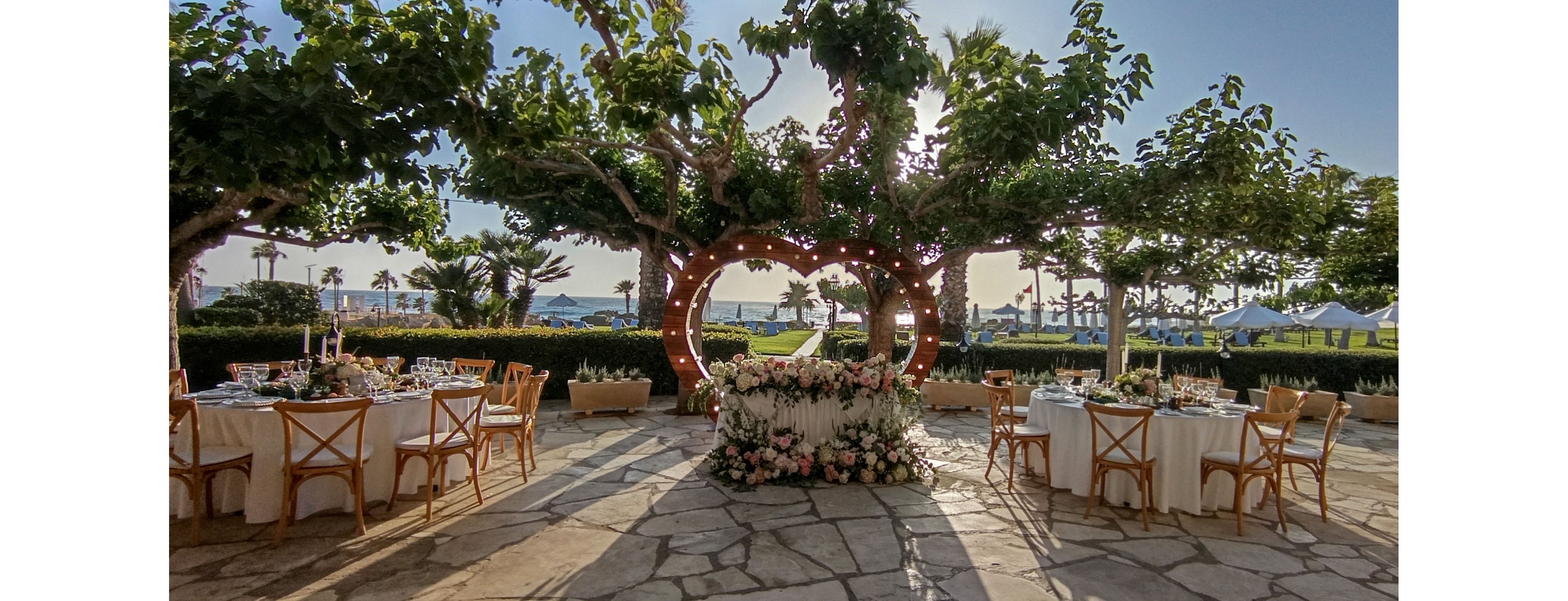 An elegant outdoor wedding reception is set beneath leafy trees with a view of the sea in the background. A circular wooden arch decorated with flowers and string lights frames the central ceremony or sweetheart table. Round tables with white linens, wooden chairs, and floral centerpieces are arranged on a stone patio. The scene is bathed in warm daylight, creating a romantic and festive atmosphere.
