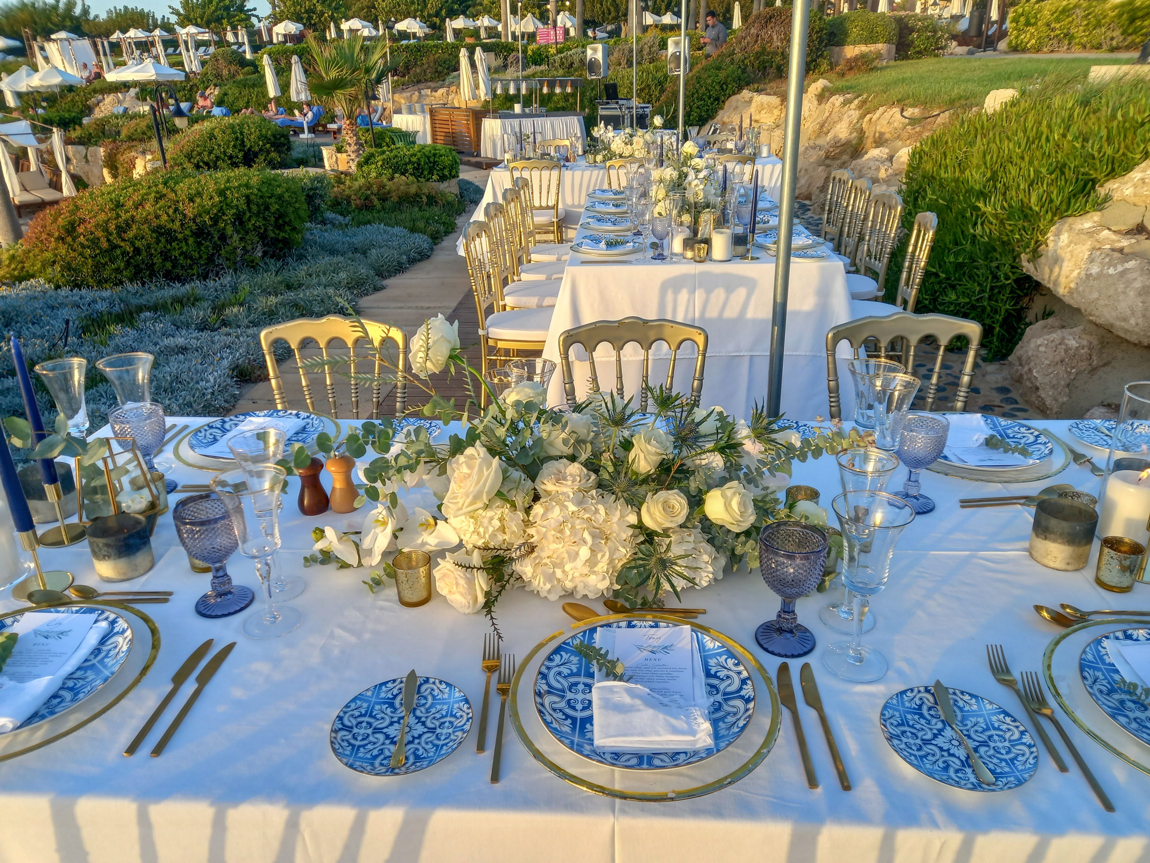 A long, elegantly set banquet table is arranged outdoors with a view of landscaped gardens and distant seating areas. The table features white linens, blue and white patterned plates, gold cutlery, and crystal glassware. Lush white floral centerpieces with greenery run along the middle of the table, creating a sophisticated and romantic atmosphere. Warm evening light enhances the refined details, suggesting a formal event or celebration.