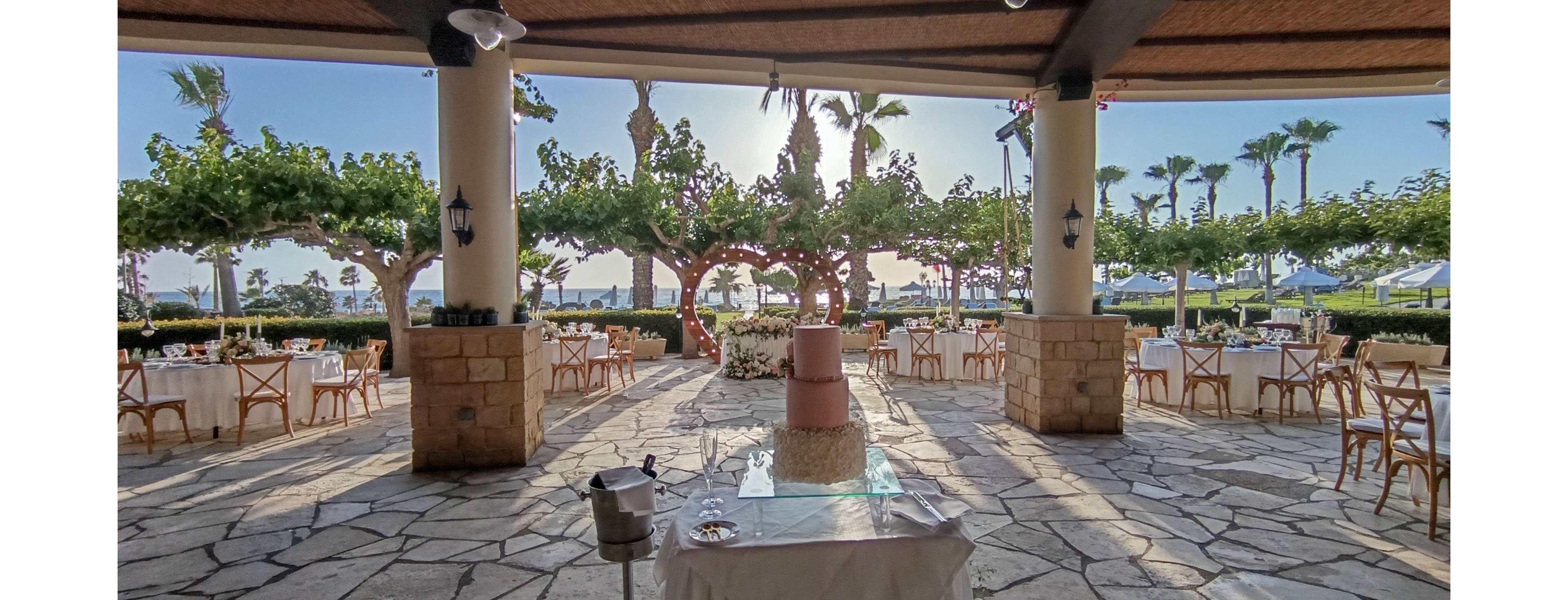 An elegant outdoor wedding reception is arranged under a covered terrace overlooking the sea. Round tables with white linens and wooden chairs are spaced around the stone patio. A multi-tiered pink cake stands on a central table, framed by columns and lush greenery. Palm trees, trimmed hedges, and distant umbrellas create a relaxed coastal atmosphere at sunset.
