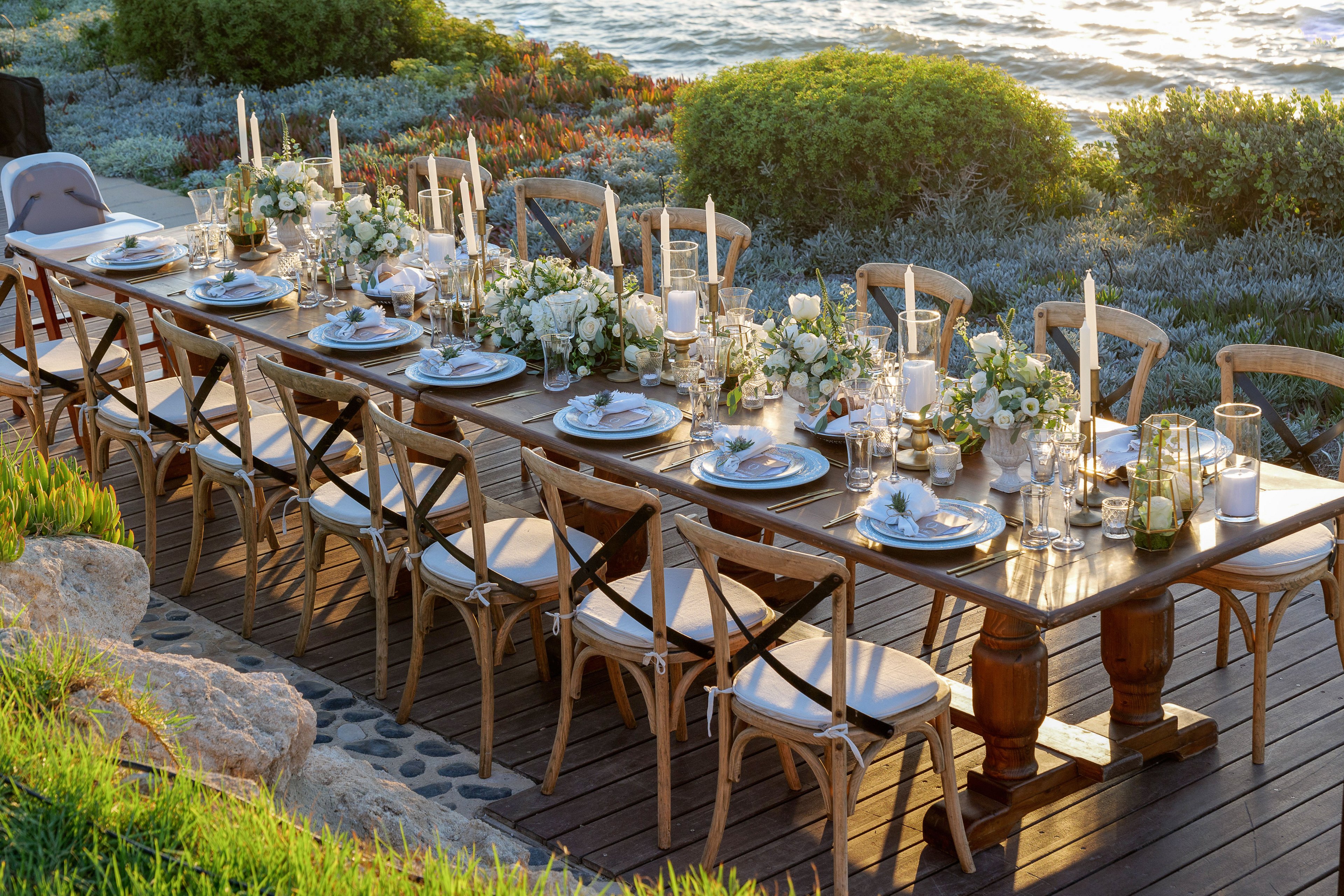 A long wooden dining table is beautifully set for an outdoor meal beside the sea. White plates, glassware, and tall candles are arranged neatly along the table. Lush white floral centerpieces and greenery run down the middle, creating a refined, romantic atmosphere. Wooden chairs with white cushions line both sides, while the ocean and coastal plants provide a scenic backdrop in warm evening light.