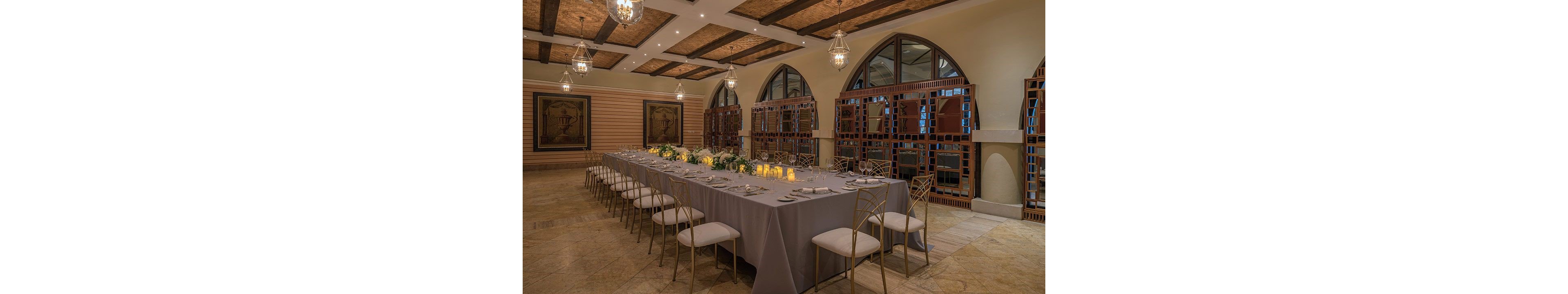A long rectangular dining table is set for a formal event in an elegant indoor banquet hall. The table is covered with a gray tablecloth and decorated with candles and greenery centerpieces. Gold-colored chairs with white cushions surround the table, ready for guests. Warm ceiling lights, hanging lanterns, arched windows, and framed wall art create a sophisticated and intimate atmosphere.