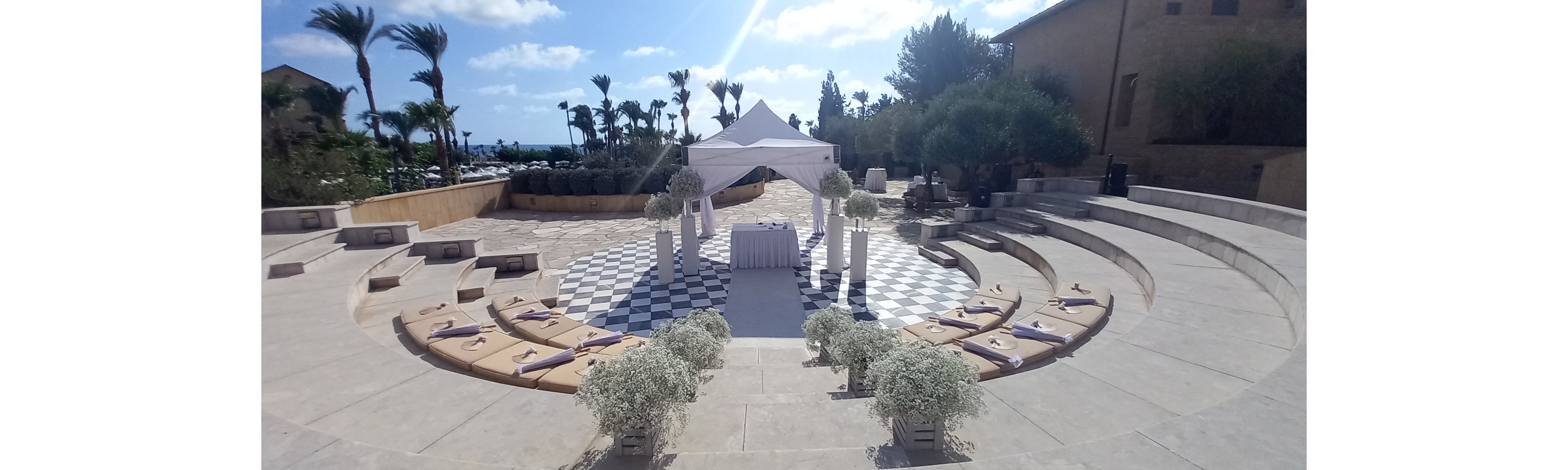 An outdoor wedding ceremony is arranged in a circular amphitheater with tiered stone seating. A white canopy stands at the center over a checkered black and white floor, framing the altar area. Cushions and rolled towels or favors are placed on the steps for guests, and white floral arrangements decorate the aisle and entrance. Palm trees, stone buildings, and a bright blue sky with scattered clouds create a sunny, coastal atmosphere.