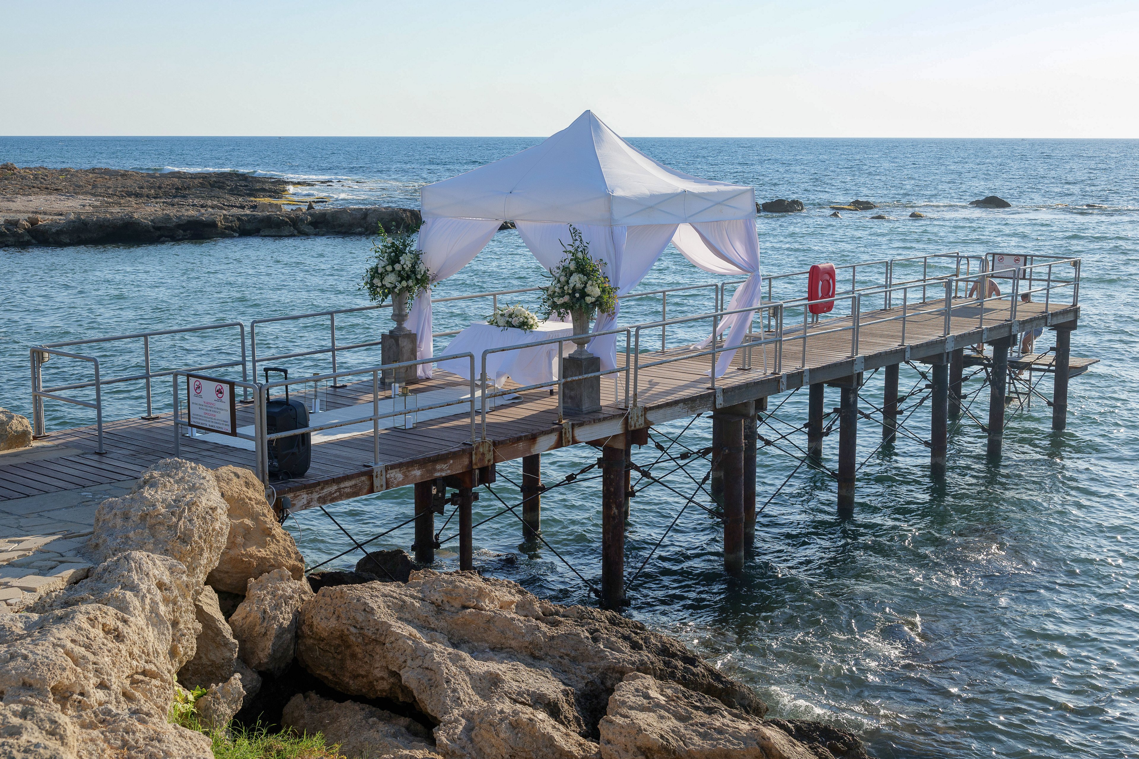 A white canopy tent is set up on a narrow pier extending over the sea. The structure is decorated with floral arrangements and a table, suggesting a romantic dinner or intimate event. Calm blue water surrounds the pier, with rocky shoreline visible in the foreground. The scene is bright and serene, evoking a peaceful coastal atmosphere.