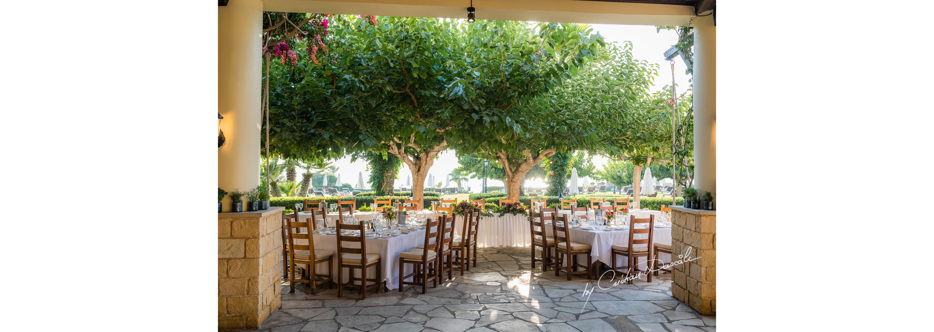An elegant outdoor reception area is arranged with round tables covered in white linens and neatly set with glassware and cutlery. Wooden chairs surround the tables, creating a warm and rustic atmosphere under a shaded terrace. Lush green trees and flowering plants frame the view, suggesting a garden or courtyard venue. The scene appears prepared for a wedding or formal event, with natural light enhancing the inviting ambiance.