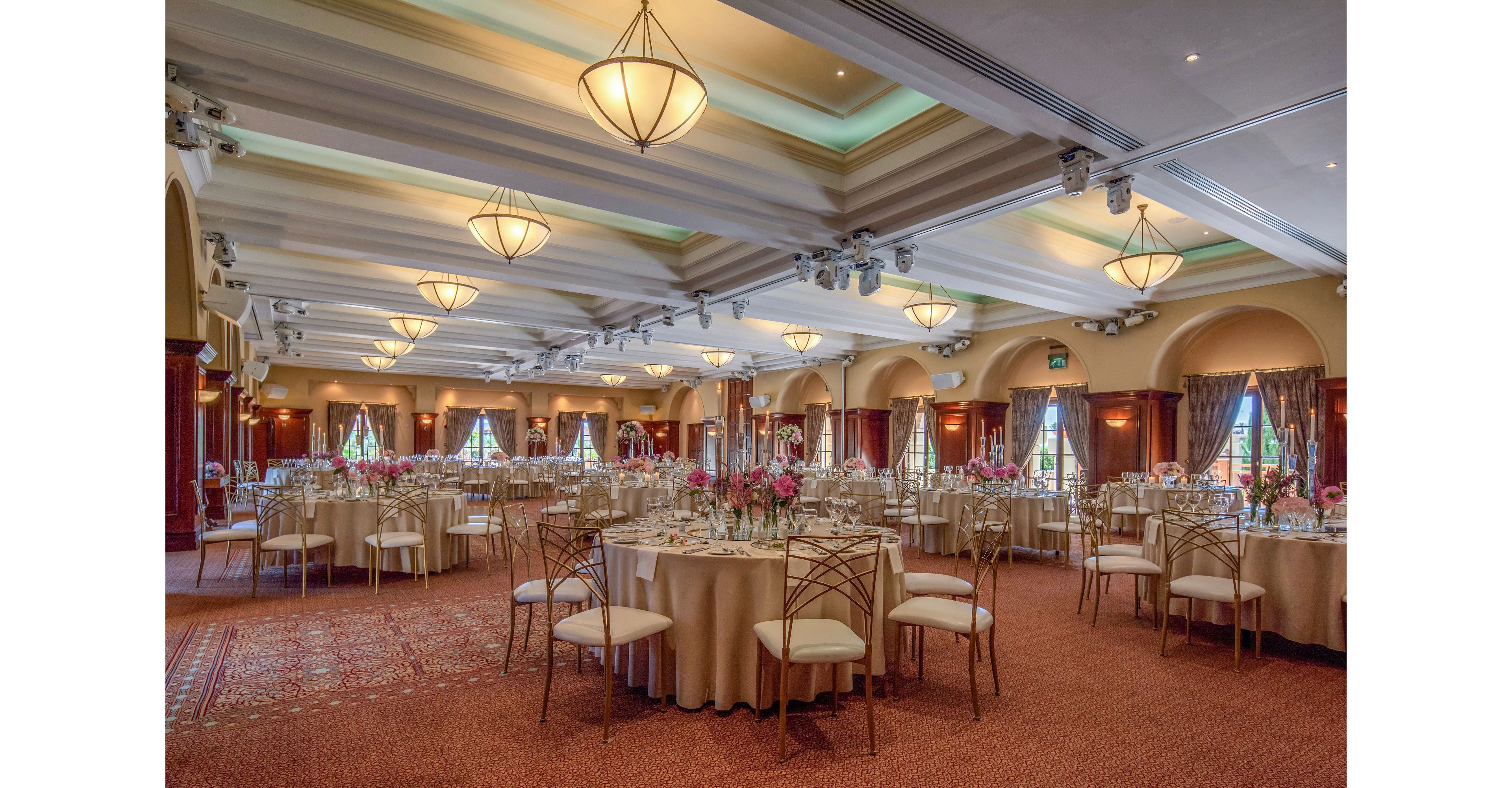 A spacious banquet hall is fully set up for a formal event with round tables covered in beige linens. Each table is arranged with glassware, plates, and floral centerpieces, creating a refined and coordinated look. Warm lighting from ceiling fixtures and wall sconces highlights the architectural details of the high ceiling and arched windows. Large windows with draped curtains allow natural light to complement the soft interior illumination, giving the room an inviting and celebratory atmosphere.