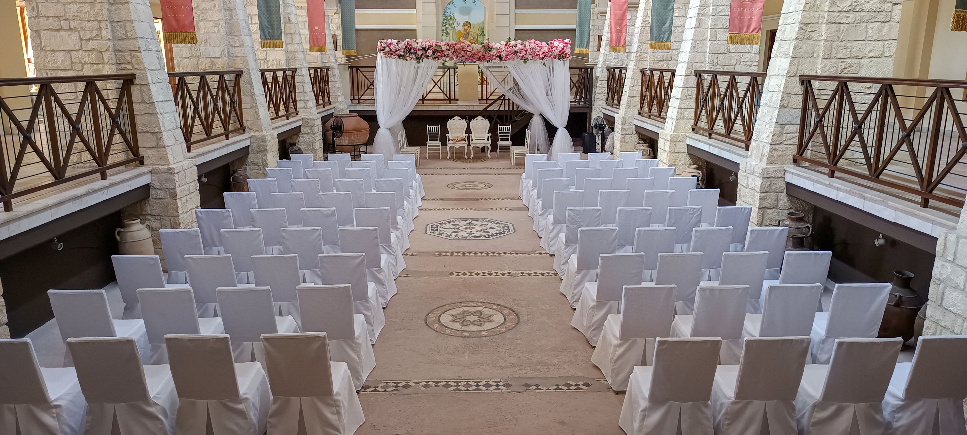 An indoor wedding ceremony venue is arranged with rows of white chair covers facing a decorated altar. The setting features stone walls, wooden railings, and a central aisle leading to a floral arch with draped white fabric. At the front, several chairs are placed beneath the arch, framed by pink and white flowers. The overall atmosphere is elegant and prepared for guests to arrive.