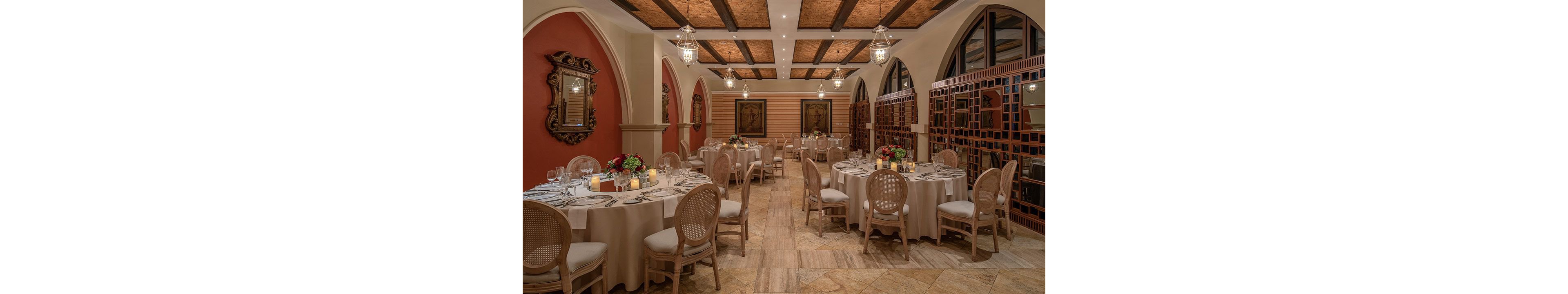 An elegant banquet room is arranged with round tables set for a formal dinner. Neutral-toned tablecloths, cushioned chairs, and floral centerpieces create a refined atmosphere. Warm lighting from hanging lanterns and candles highlights the arched doorways, decorative mirrors, and framed artwork on the walls. The overall setting suggests a sophisticated event or private dining experience in a classic, upscale venue.