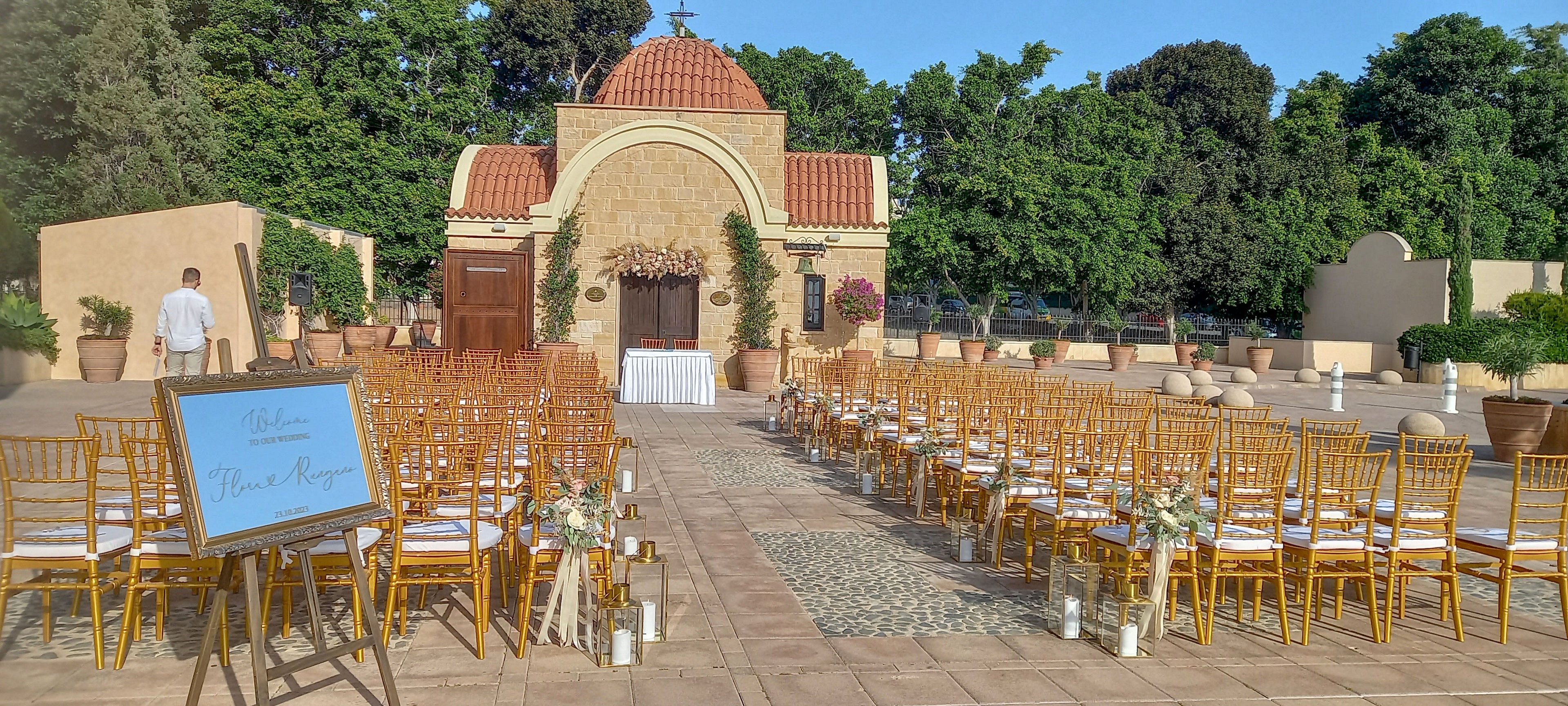 An outdoor wedding ceremony is arranged in a Mediterranean-style courtyard facing a small chapel with a red-tiled dome. Rows of gold chairs with white cushions line a central aisle decorated with white flowers and glass candle holders. A large welcome sign on an easel stands to the left, and a single person walks near the seating area. Warm sunlight and lush green trees in the background create a bright, festive atmosphere.