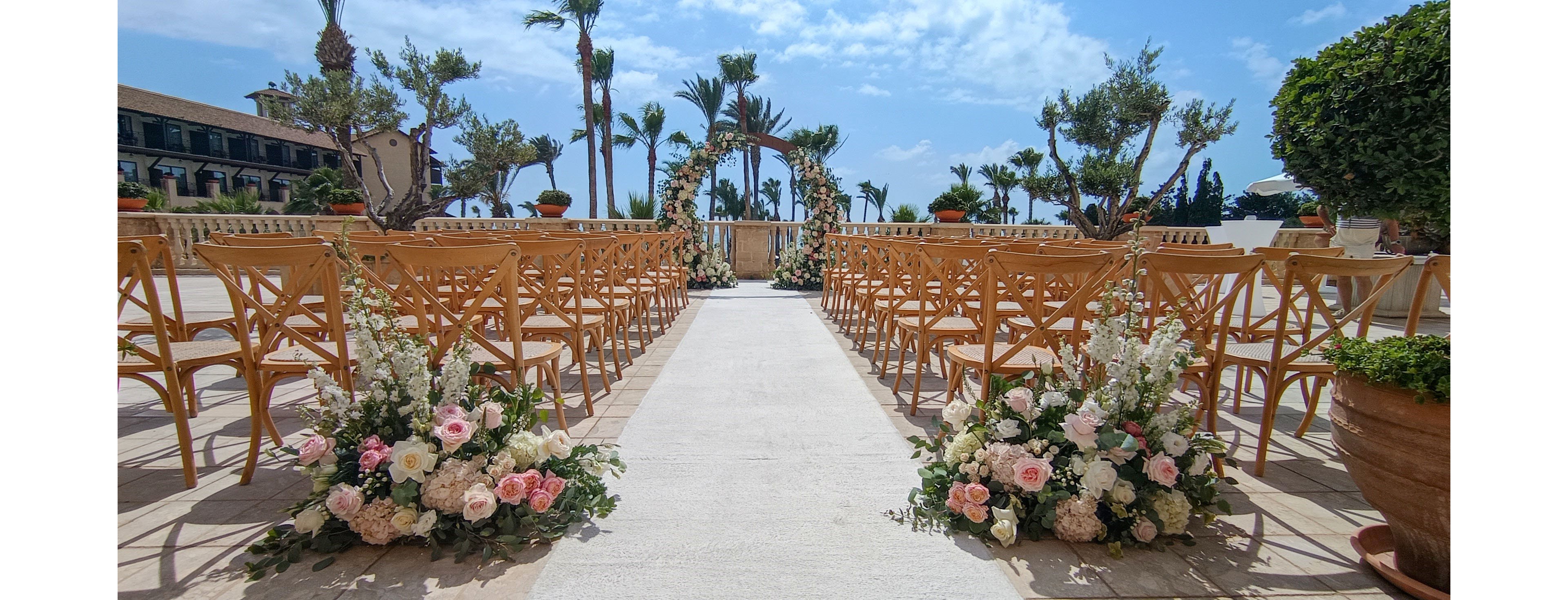 An elegant outdoor wedding ceremony setup features rows of wooden chairs facing a floral arch. The aisle is lined with lush arrangements of white and blush pink flowers at the entrance. Palm trees and manicured greenery frame the bright, sunny setting under a blue sky with scattered clouds. The scene conveys a romantic, coastal or Mediterranean ambiance ready for guests to arrive.