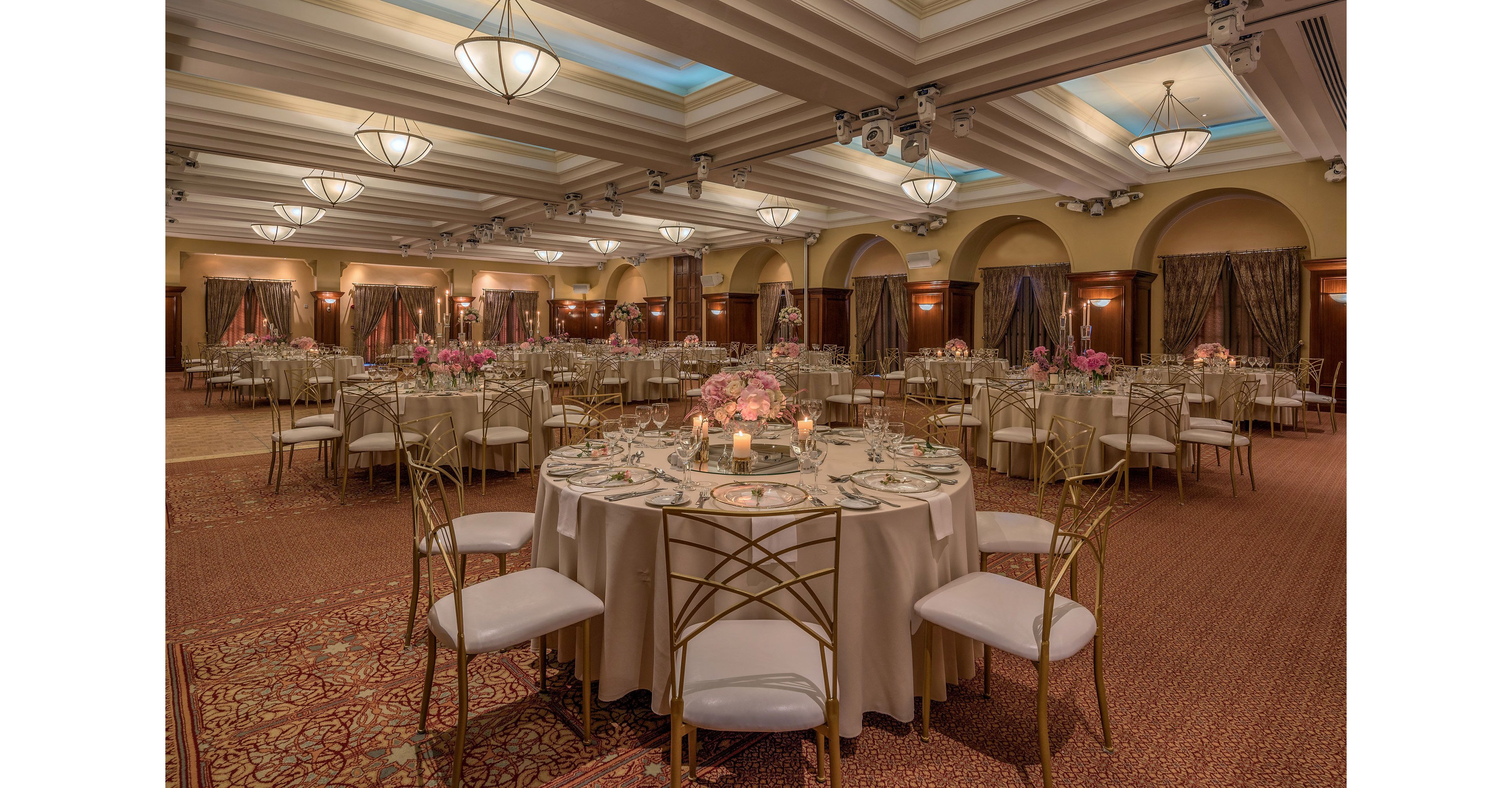 A large banquet hall is arranged with round tables covered in beige linens and surrounded by gold chairs with white cushions. Each table is set with glassware, plates, and silverware, and decorated with pink floral centerpieces and candles. Warm ceiling lights and chandeliers illuminate the room, highlighting the patterned carpet and draped curtains along the walls. The overall atmosphere suggests a formal event such as a wedding reception or gala dinner.