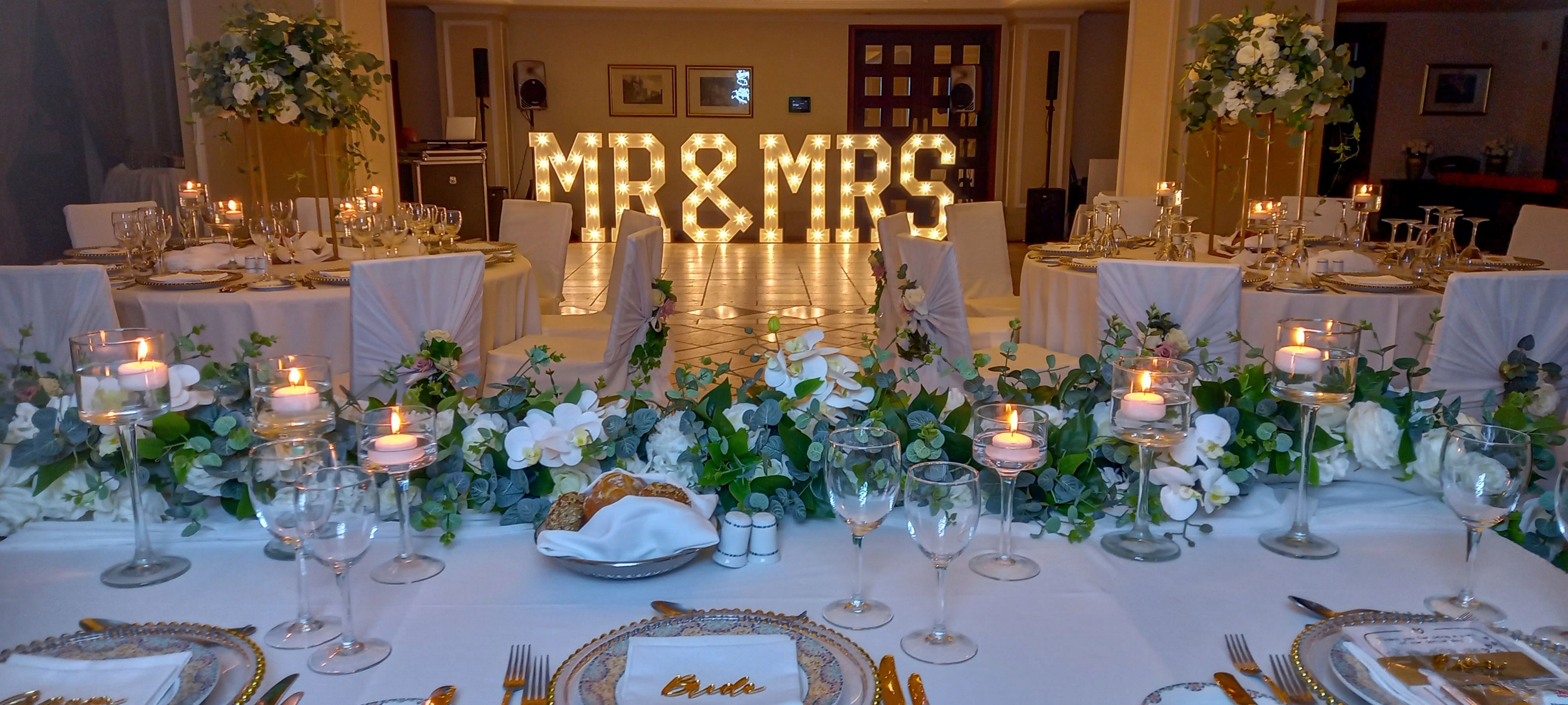 A luxurious wedding reception table is set with white linens, crystal glassware, and gold-accented plates. Lush green and white floral arrangements run along the center, interspersed with numerous glowing candles. In the background, a large illuminated MR & MRS sign creates a romantic focal point in the hall. The warm lighting and refined decor suggest an intimate, upscale celebration.
