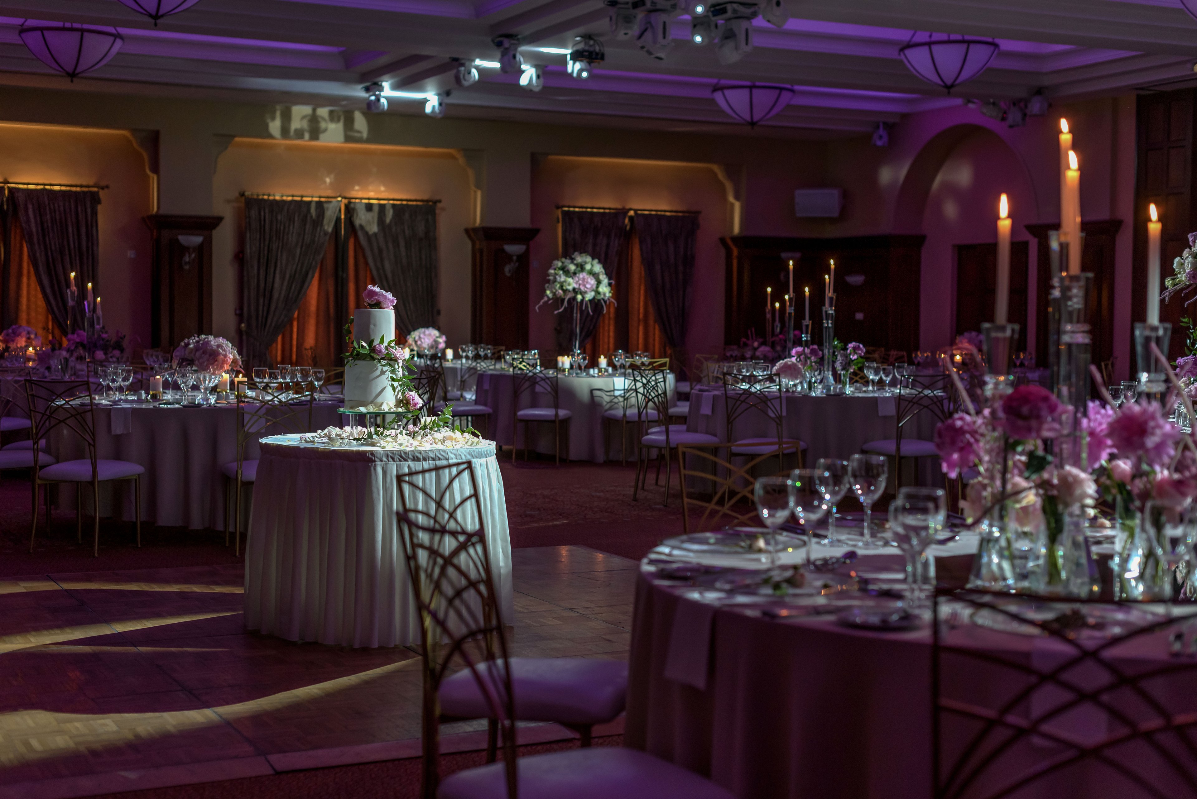 A softly lit wedding reception hall is arranged with round tables and a central multi-tiered cake. The room features neatly set glassware, floral centerpieces, and candles creating a romantic atmosphere. Purple and pink lighting casts gentle highlights across the tables and chairs. Draped curtains and classic architectural details frame the sophisticated indoor setting.