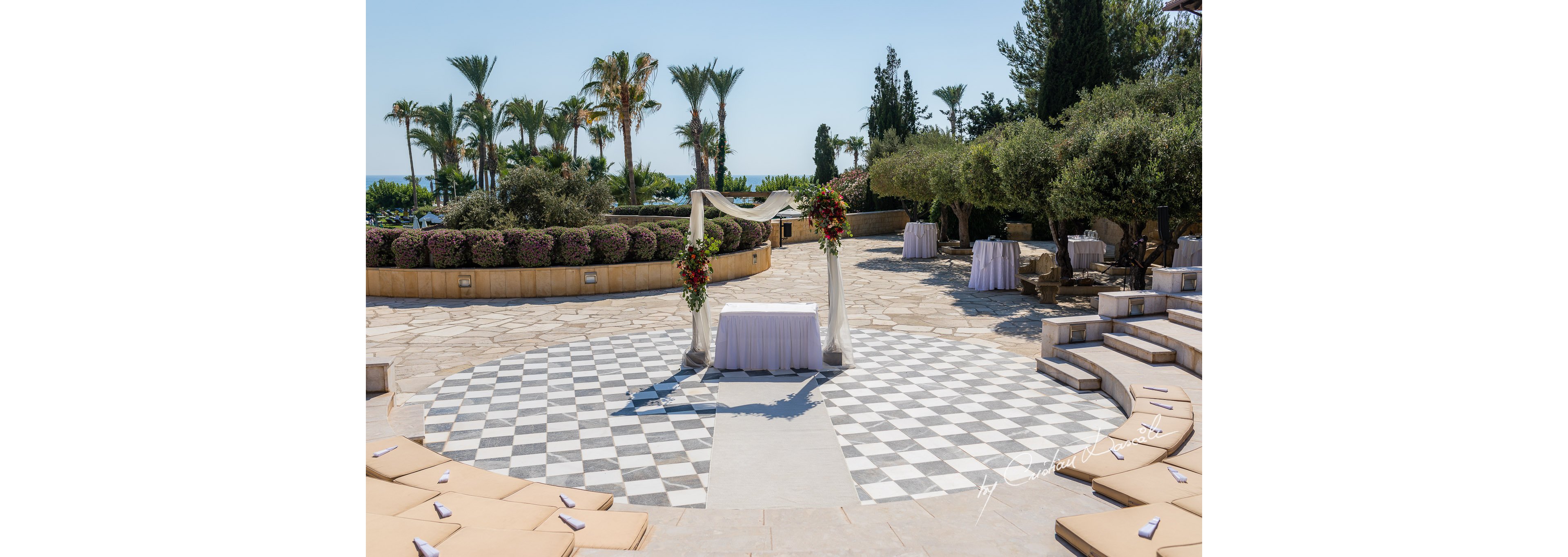 An elegant outdoor wedding ceremony area is arranged on a circular checkerboard stone plaza. A simple white arch with floral arrangements frames a small table draped in white fabric. Curved seating steps with beige cushions surround the central space, creating an intimate amphitheater feel. Palm trees, flowering shrubs, and a distant view of the sea provide a scenic coastal backdrop under a clear blue sky.