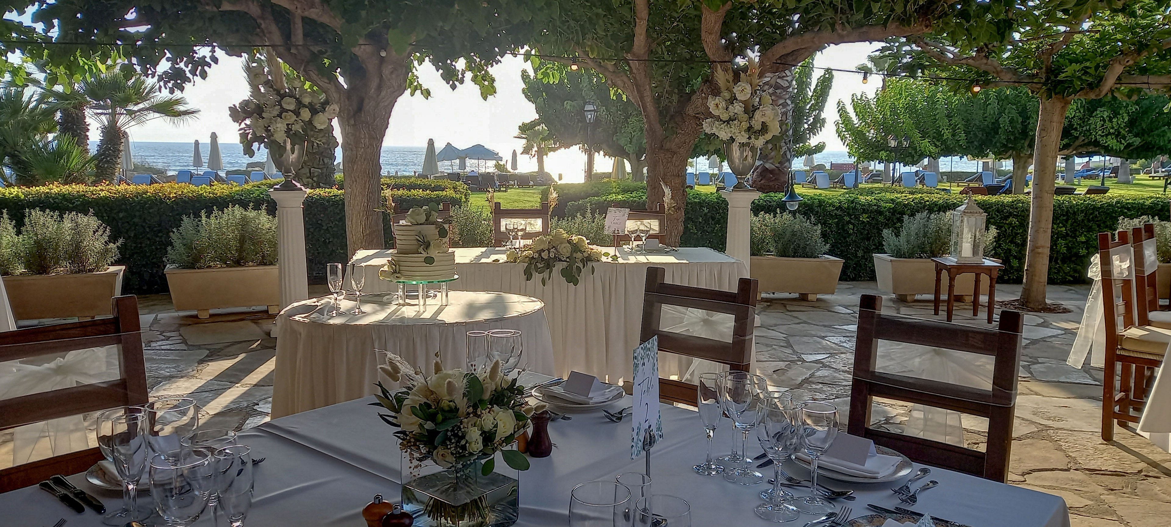 An elegant outdoor wedding reception is set beneath leafy trees overlooking the sea. White-clothed tables are arranged with glassware, cutlery, and floral centerpieces, creating a refined dining atmosphere. Wooden chairs surround the tables on a stone patio, framed by manicured hedges and potted plants. The warm sunlight filters through the trees, adding a romantic glow to the coastal setting.