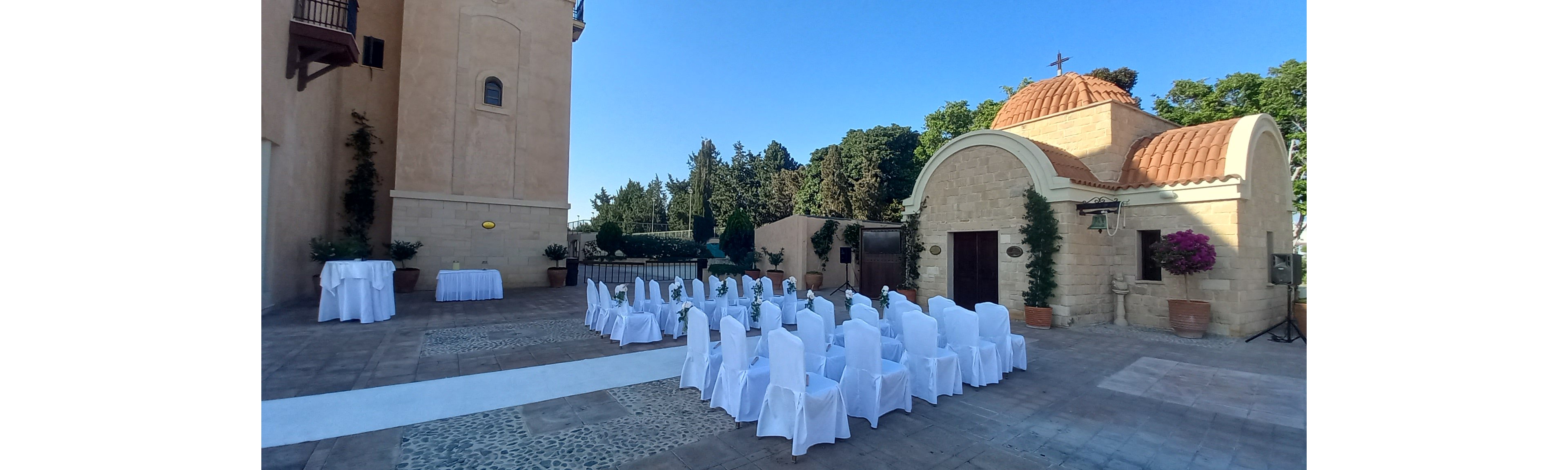 An outdoor wedding ceremony is arranged in front of a small chapel with a domed roof and cross. Rows of white-covered chairs line a white aisle runner on a stone courtyard. The surrounding buildings feature warm beige walls, arched doorways, and potted plants. Bright daylight and clear blue sky create a serene and romantic atmosphere.