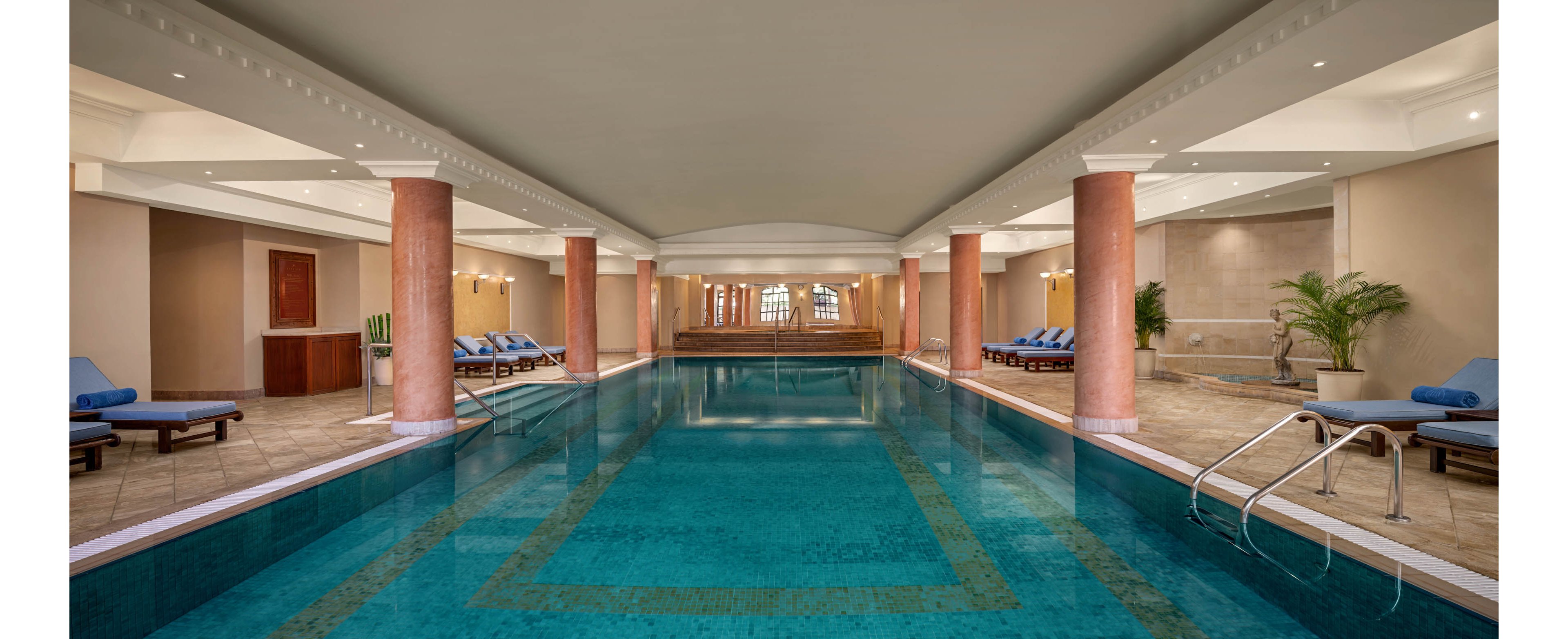 A long indoor swimming pool stretches between rows of pink marble columns in a hotel or spa setting. Blue lounge chairs with towels line both sides of the pool on a tiled floor. Warm neutral walls, recessed ceiling lights, and potted plants create a calm, upscale atmosphere. Stainless steel ladders and clear reflections in the water emphasize cleanliness and relaxation.