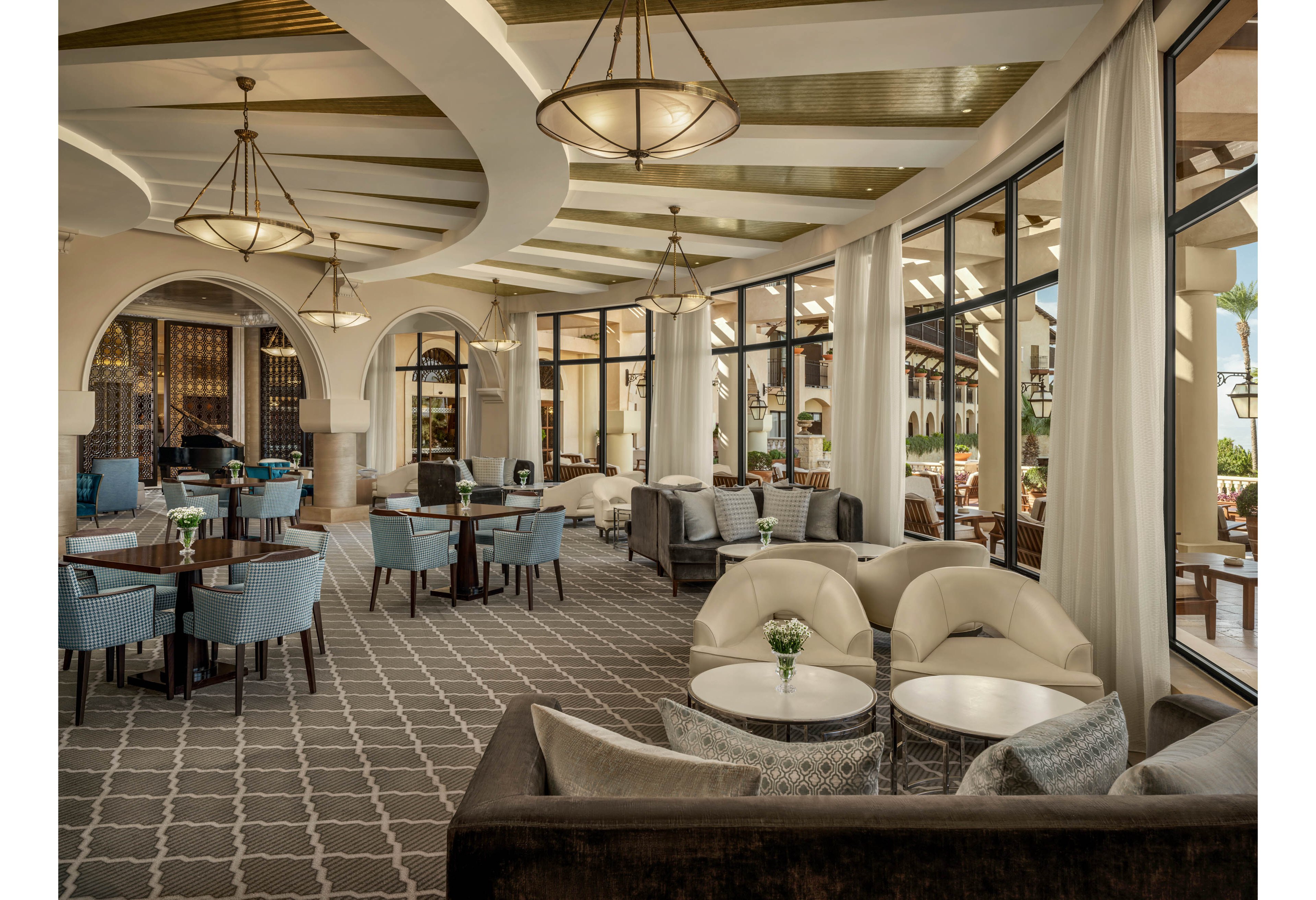 A spacious hotel lounge features a mix of armchairs, sofas, and dining tables arranged in an open-plan setting. Large floor-to-ceiling windows allow natural light to fill the room and provide views of an outdoor terrace. Neutral tones with blue accents, patterned carpeting, and arched architectural details create a sophisticated atmosphere. Small floral arrangements on the tables add a refined decorative touch.