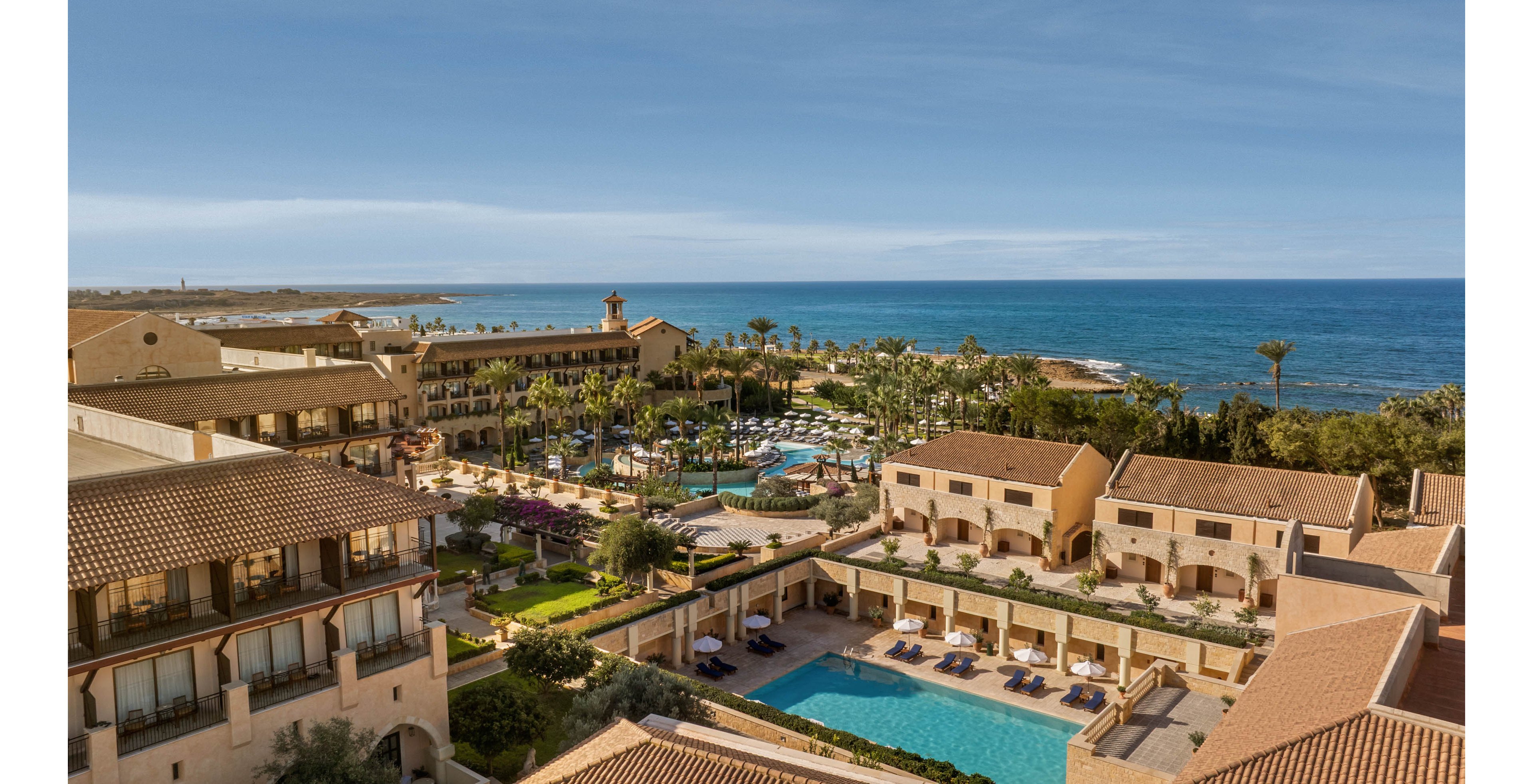 A large seaside resort complex stretches along the coastline, featuring multiple tan-colored buildings with tiled roofs. In the center, several swimming pools are surrounded by palm trees, sun loungers, and umbrellas, creating a relaxing vacation atmosphere. The calm blue ocean extends to the horizon under a clear sky, emphasizing the coastal setting. The overall scene suggests a sunny, tranquil holiday destination with extensive amenities.