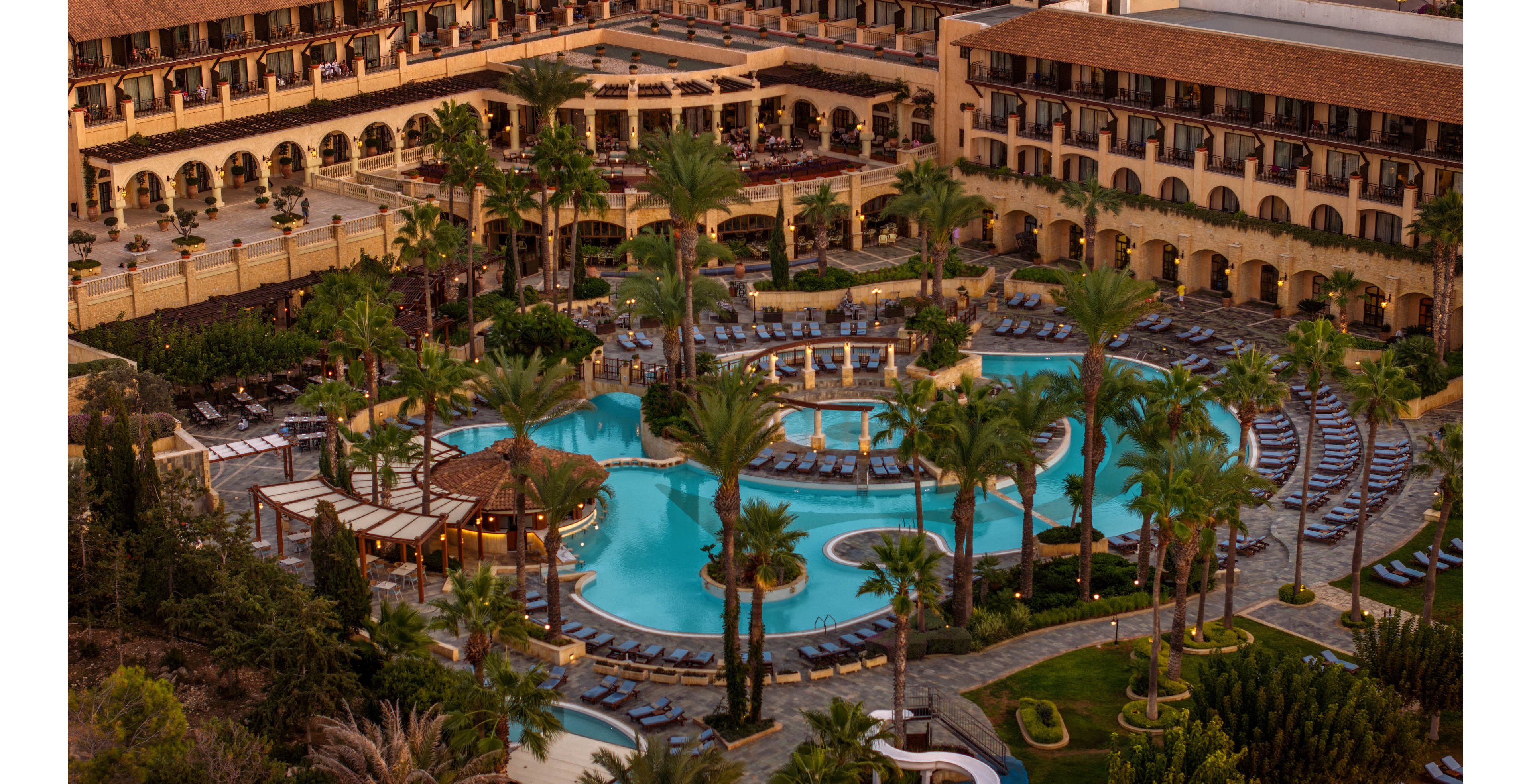 An expansive luxury resort features a large freeform swimming pool surrounded by palm trees and sun loungers. Multiple levels of terraced buildings with arches and balconies overlook the central pool area. The scene is captured at sunset or golden hour, casting warm light across the water and architecture. Curved pathways, cabanas, and landscaped gardens add to the tropical vacation atmosphere.