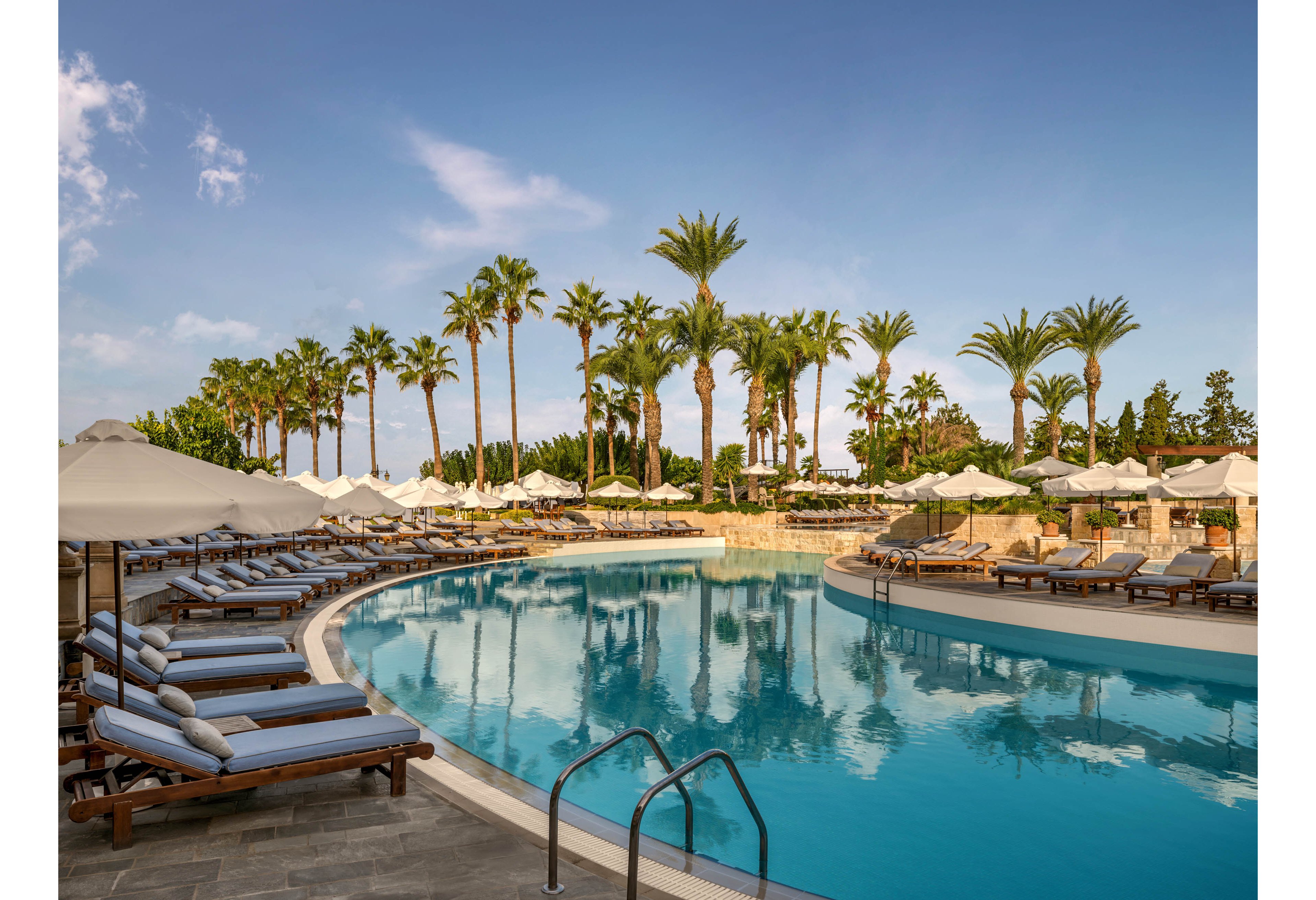 A large curved swimming pool is surrounded by neatly arranged lounge chairs and umbrellas at a luxury resort. Tall palm trees line the background, reflecting in the calm blue water under a clear sky. Cushioned sunbeds and shaded seating areas suggest a relaxing vacation atmosphere. The scene appears quiet and serene, with no people visible around the pool.