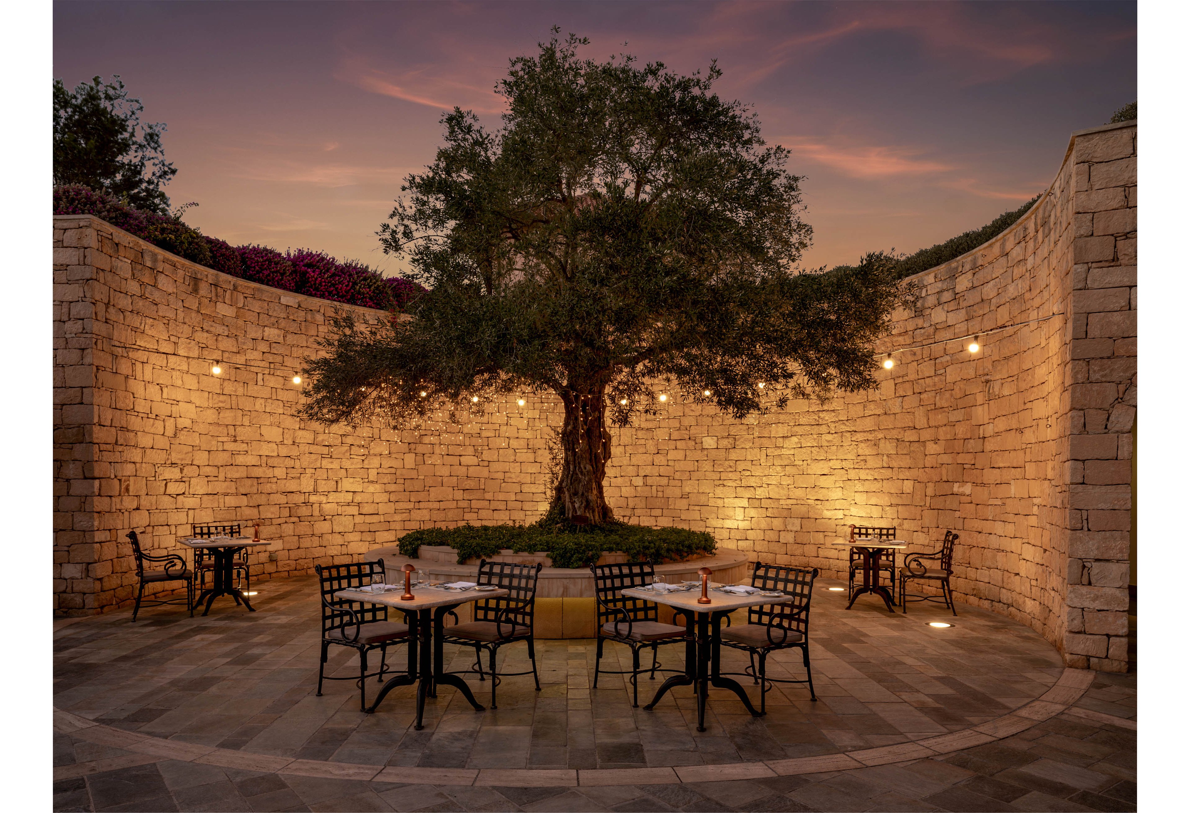 An elegant outdoor courtyard features a large tree at its center, dramatically lit against curved stone walls. Several small round tables with metal chairs are arranged around the tree, each set for an intimate meal. Warm string lights and ground lighting create a cozy, inviting glow as the sky transitions into sunset. The scene suggests a tranquil, romantic dining experience in a secluded garden setting.