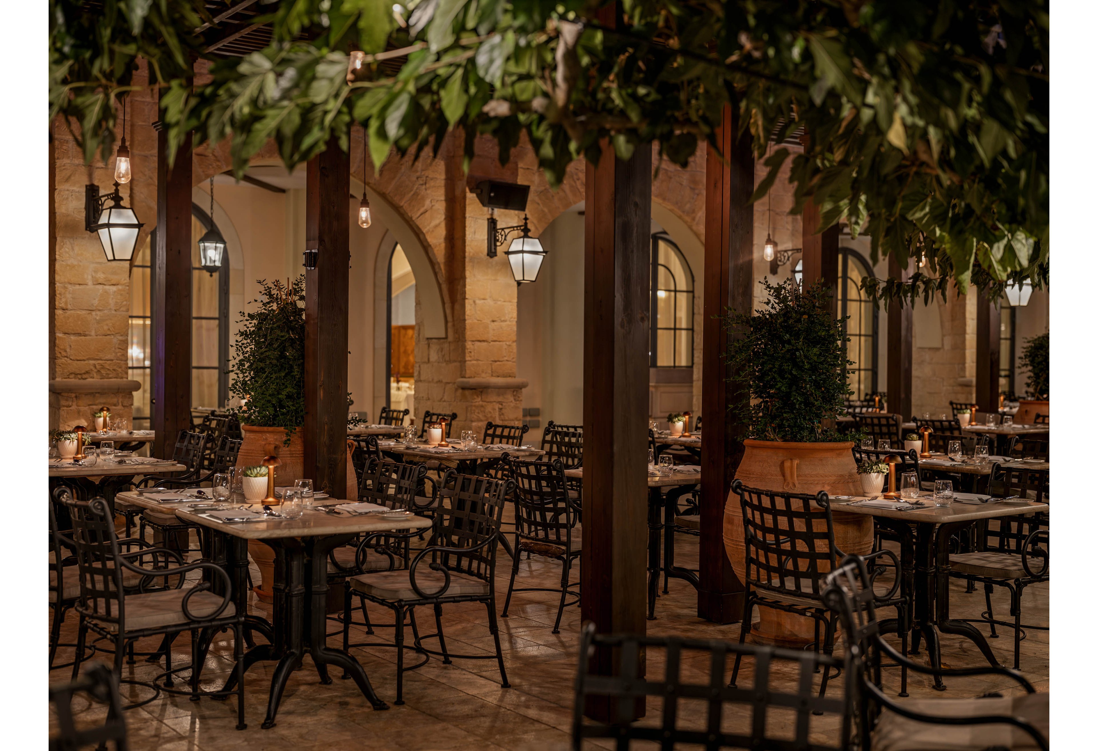 An elegant outdoor restaurant courtyard is softly lit in the evening. Empty wrought iron tables and chairs are arranged beneath leafy trees and hanging lanterns. Warm light from candles and wall sconces reflects off the stone arches and tiled floor. The scene feels intimate and inviting, ideal for a romantic dinner or special event.
