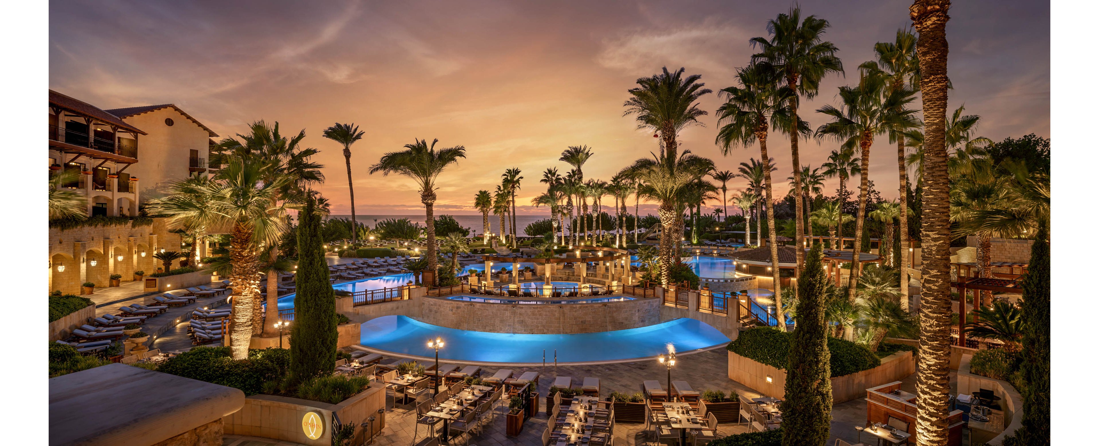 A luxurious resort features multiple tiered swimming pools surrounded by palm trees at dusk. Outdoor dining tables and lounge chairs are arranged neatly around the illuminated pools. Warm golden lighting from the buildings contrasts with the deep blue water and colorful sunset sky. The scene suggests a tranquil, upscale vacation atmosphere by the sea.