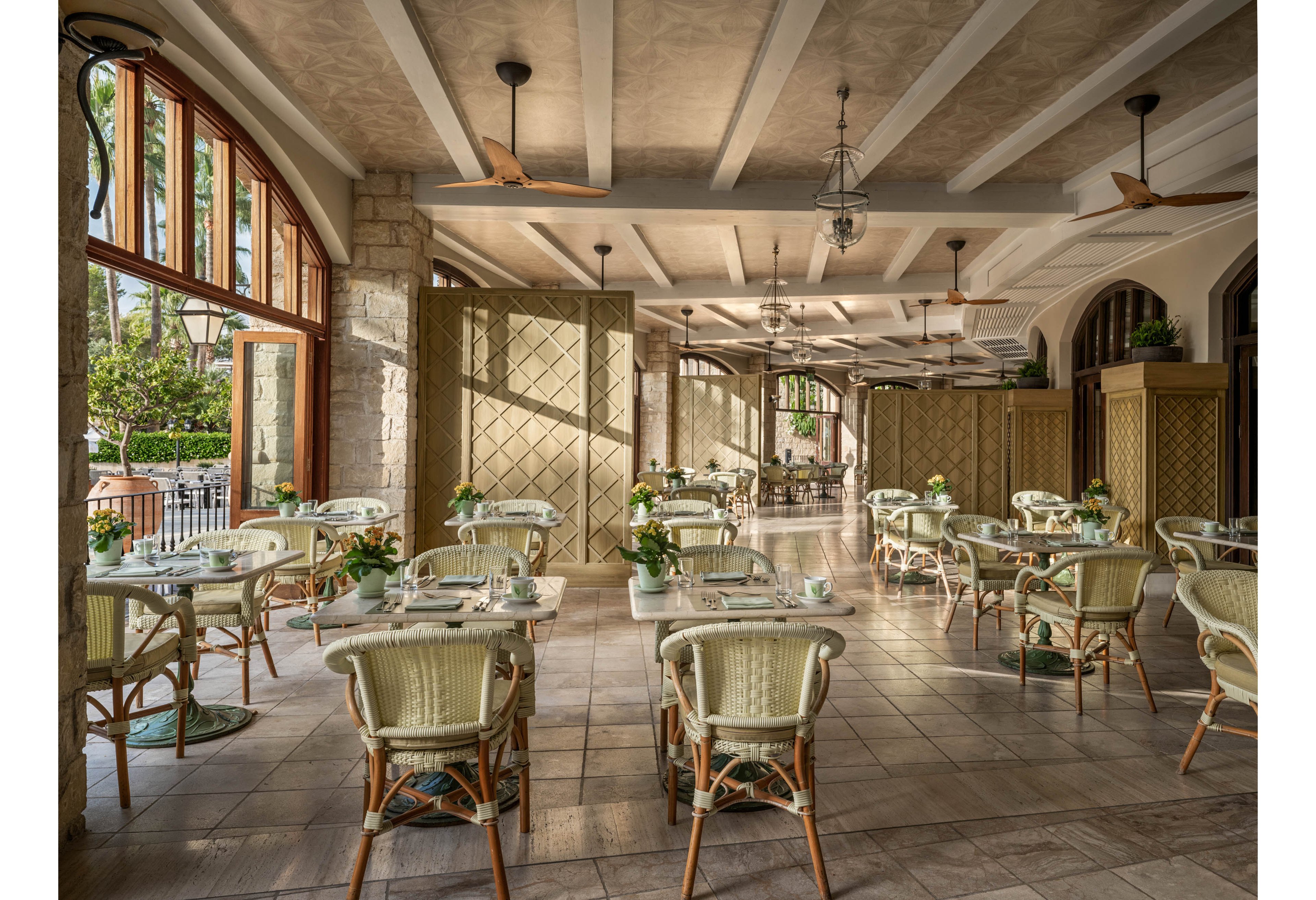An elegant open-air restaurant features light wicker chairs and small round tables arranged neatly across a tiled floor. Large arched windows and open doors let in abundant natural light and views of greenery outside. Ceiling fans and exposed beams add to the airy, coastal-inspired design. Potted plants on each table contribute to a fresh, inviting atmosphere in the empty dining space.