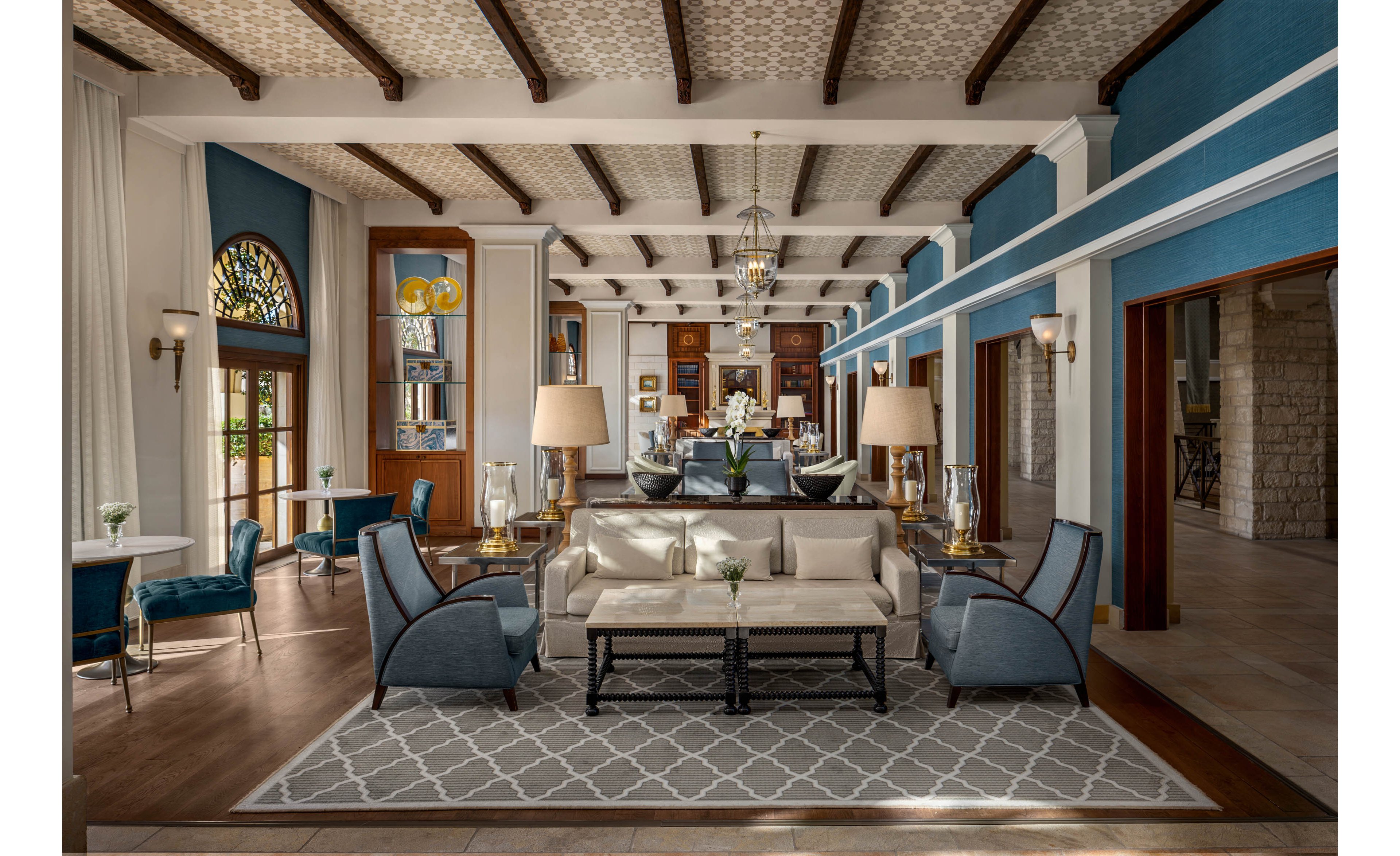 A sophisticated hotel lobby features a central seating area with cream sofas and blue armchairs arranged around a patterned rug. The space is framed by tall white columns, wooden ceiling beams, and large windows with sheer curtains that allow natural light to filter in. Warm table lamps, decorative flowers, and framed artwork add a refined, welcoming atmosphere. In the background, additional seating and a reception or lounge area extend the sense of depth and luxury.