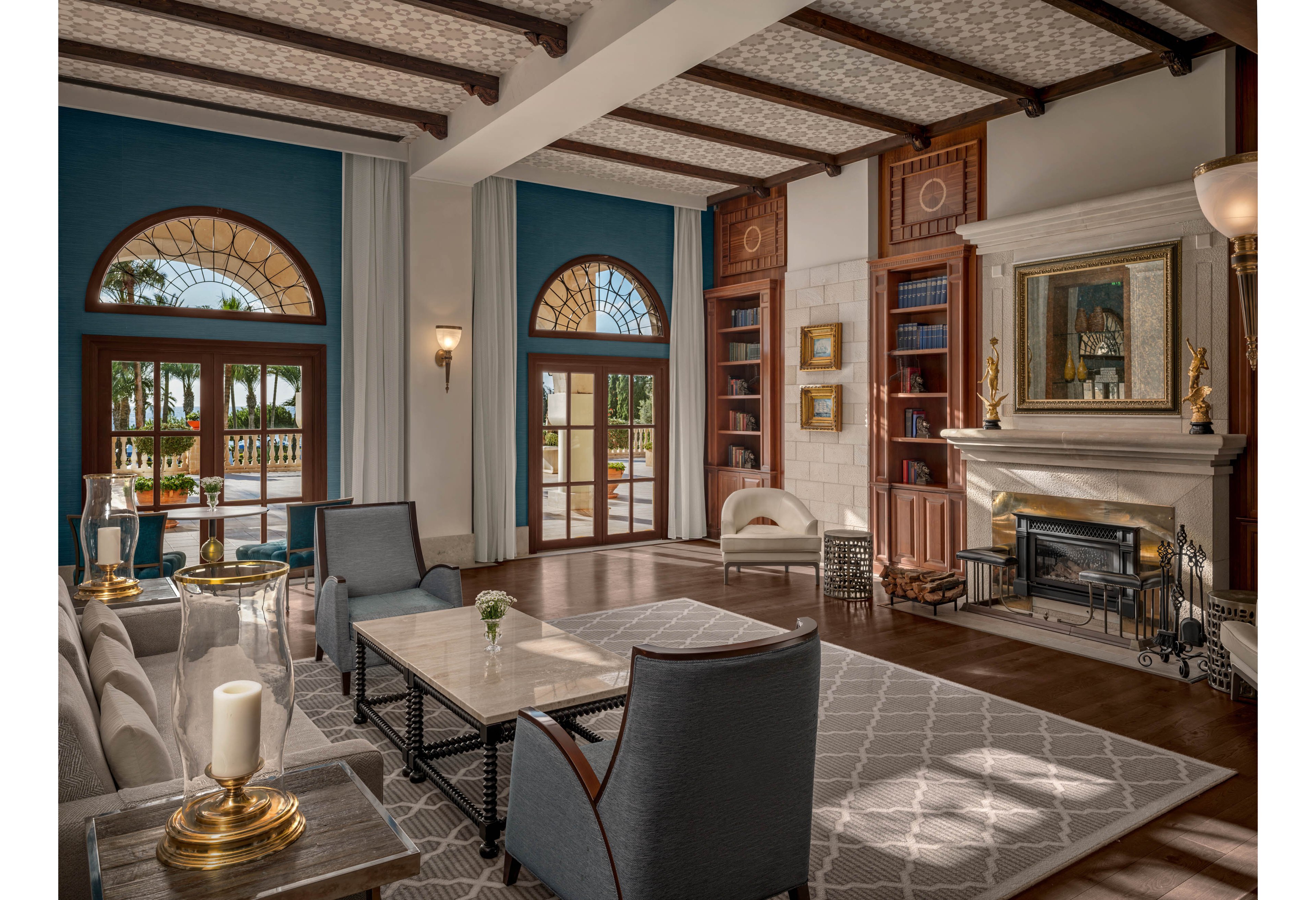 A spacious, elegant lounge features plush seating arranged around a central coffee table and a decorative fireplace. Large arched windows and glass doors allow natural light to fill the room and reveal palm trees outside. Built-in wooden bookshelves, framed artwork, and sculptural decor add a refined, classic touch. Soft neutral tones with blue accents and patterned rugs create a warm, inviting atmosphere suitable for a hotel or upscale residence.